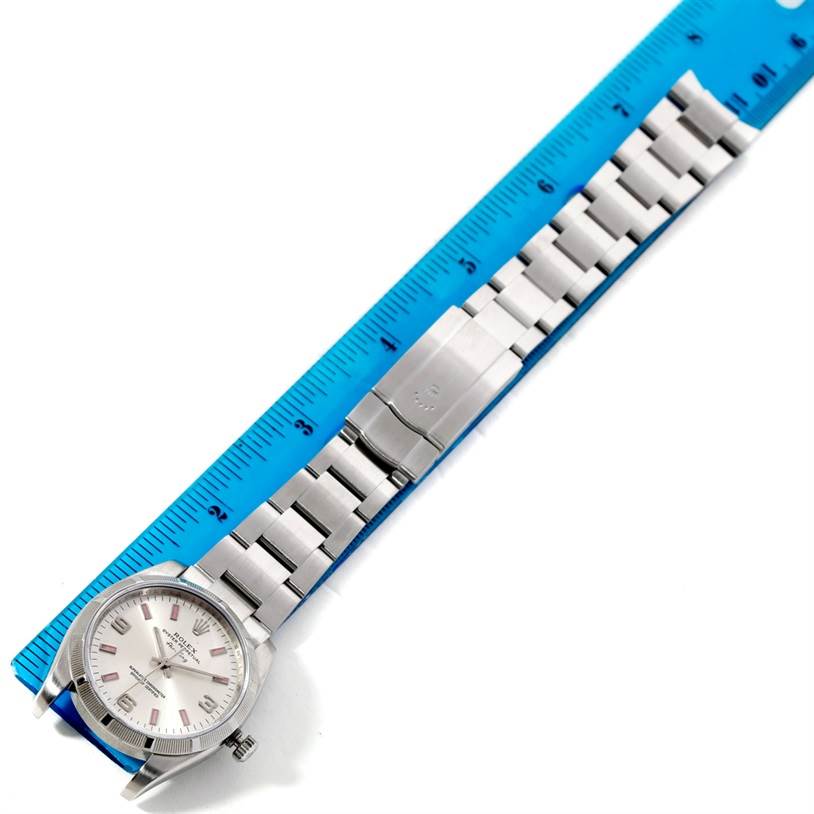 The image shows a top-down view of a Rolex Air-King 114200 Men's Stainless Steel Silver Dial 114200 Men's Stainless Steel Silver Dial watch with its silver bracelet aligned alongside a blue measuring ruler.