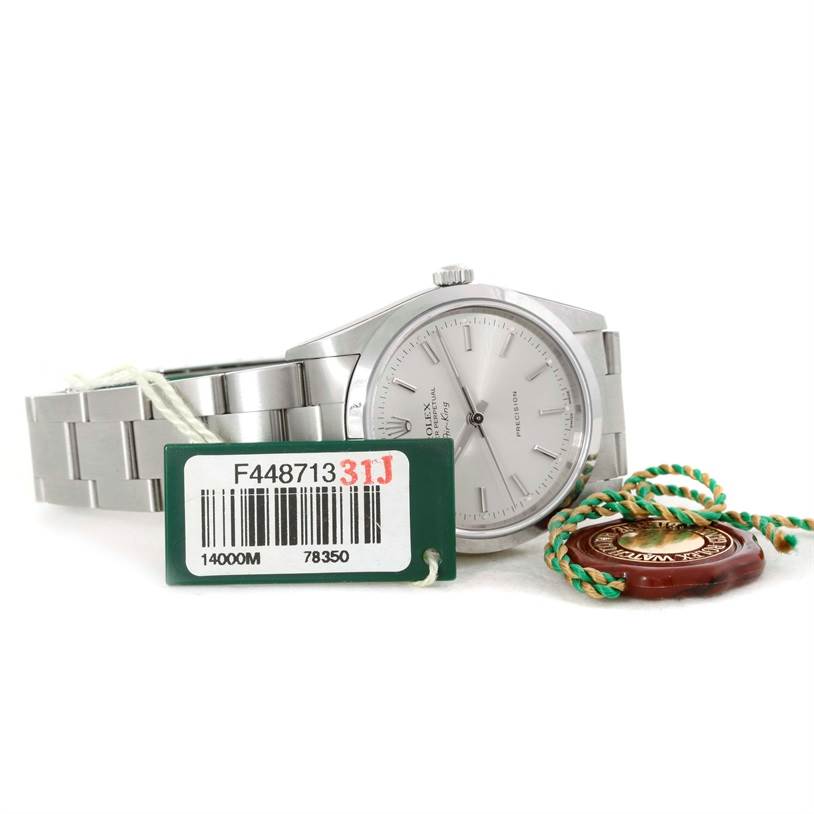 The Rolex Air-King 14000 Men's Stainless Steel Silver Dial 14000 Men's Stainless Steel Silver Dial watch is shown from a front angle, accompanied by a tag and a green string with a medallion.