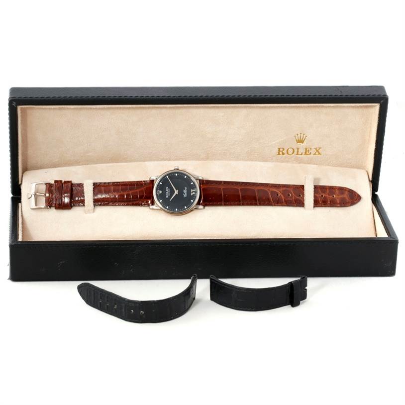 The Rolex Cellini 5116 Men's White Gold Black Dial 5116 Men's White Gold Black Dial watch is shown from a top-down angle inside its box, with additional black straps placed outside.