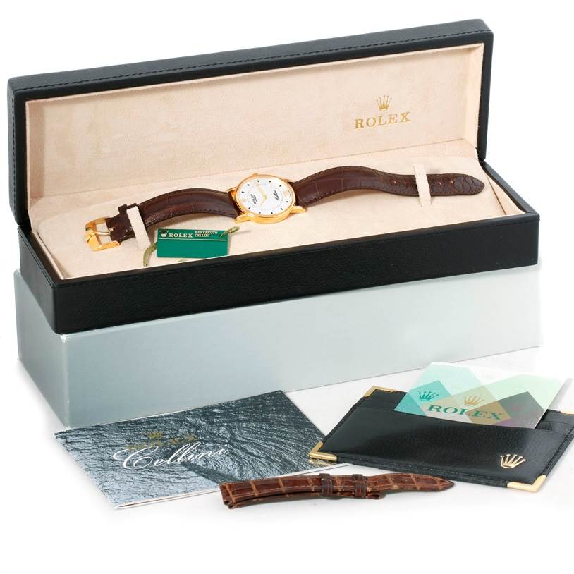 The Rolex Cellini 5115 Men
s Yellow Gold Silver Dial 5115 Men
s Yellow Gold Silver Dial watch is shown at an angle inside its box, revealing the dial, strap, warranty card, manual, extra strap, and a leather wallet.