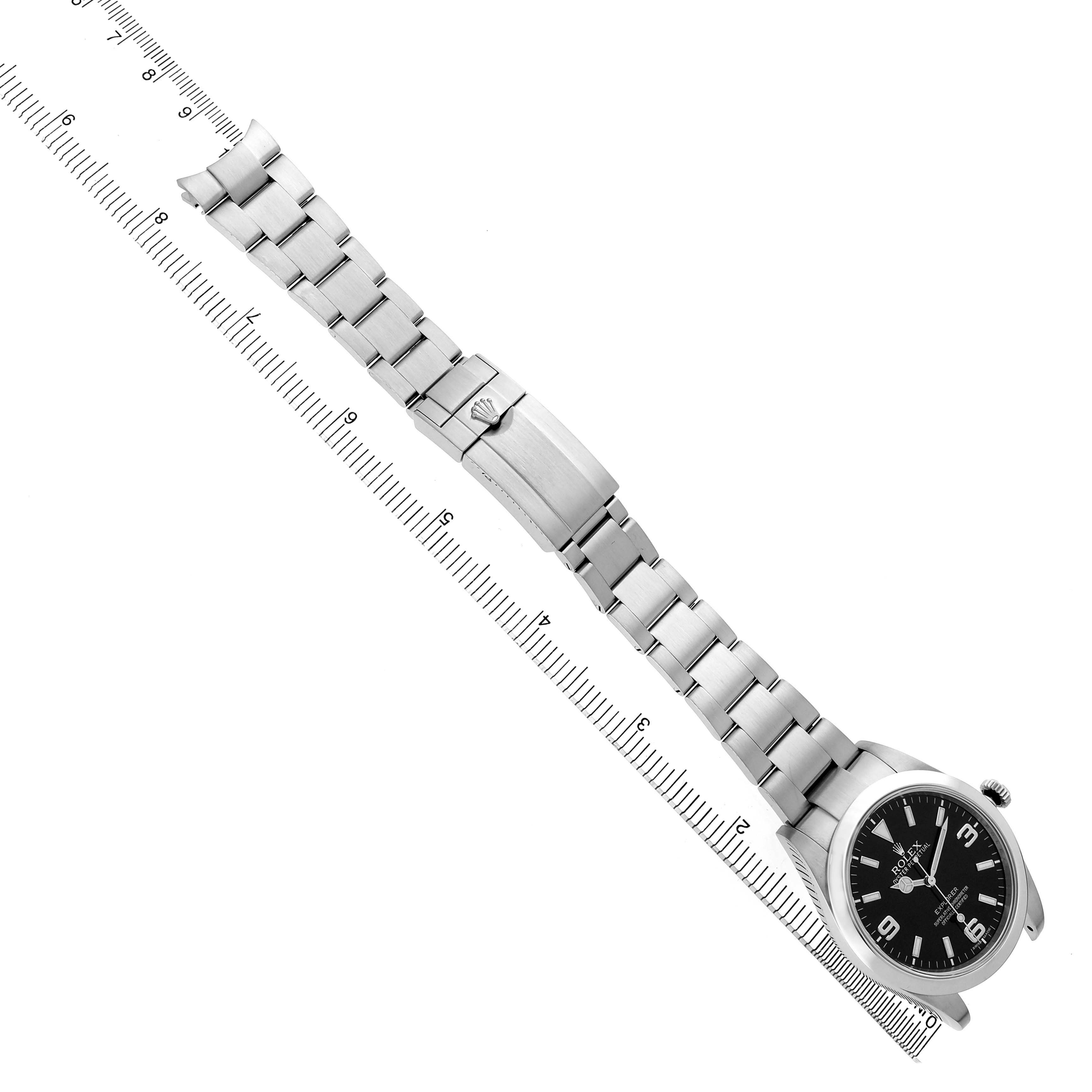 The image shows a Rolex Explorer 214270 Men's Stainless Steel Black Dial watch laid flat with its full metal bracelet extended and a clasp, against a ruler for scale.