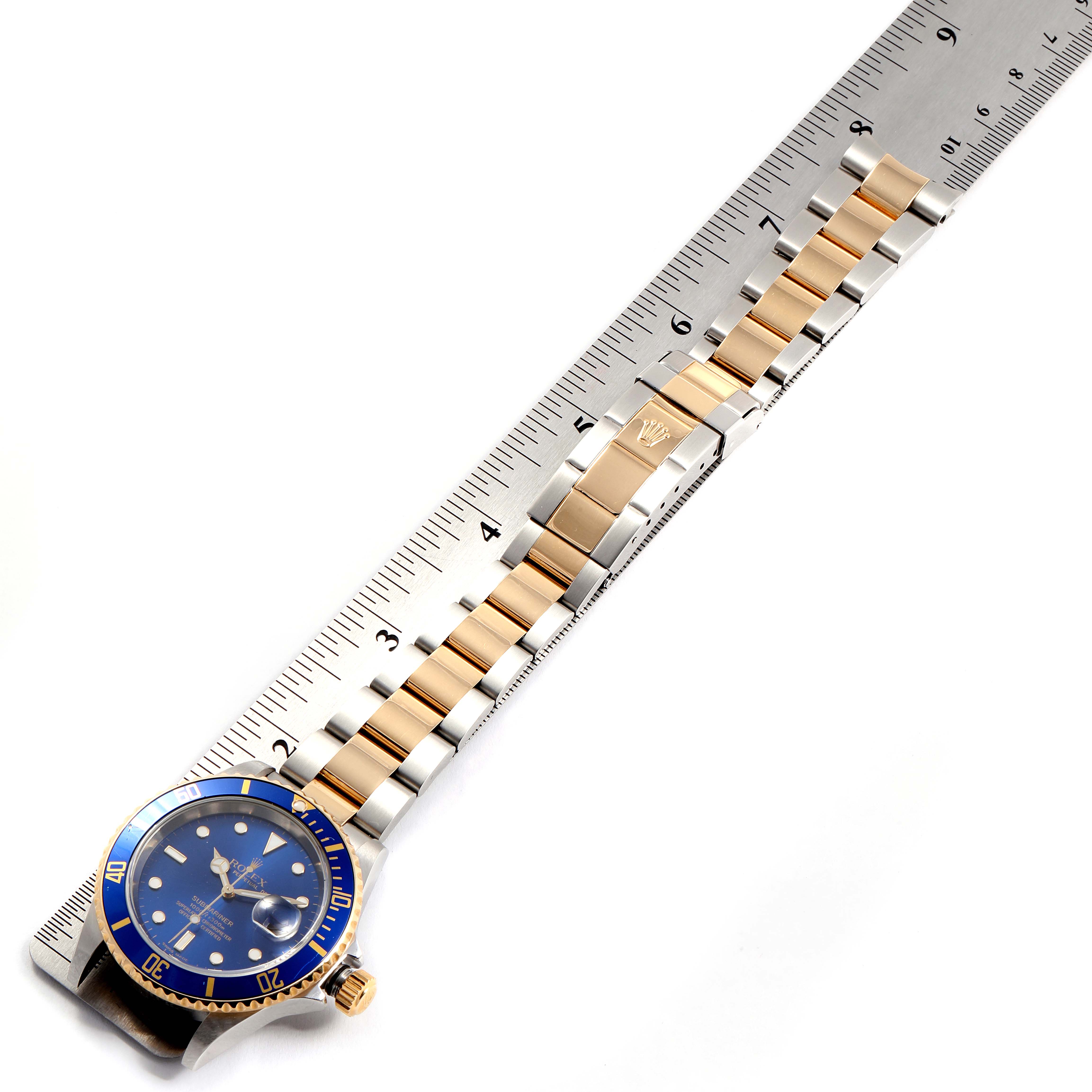The image shows a Rolex Submariner 16613 Men
s Steel and Gold (two tone) Blue Dial 16613 Men
s Steel and Gold (two tone) Blue Dial watch from an overhead angle, highlighting the blue bezel, face, and the two-tone bracelet laid on a ruler.