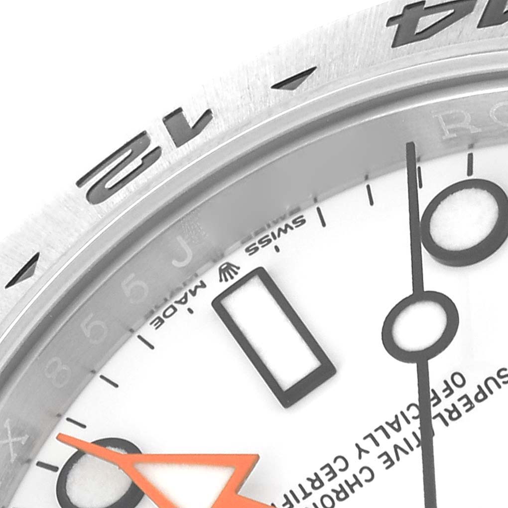 This close-up image of the Rolex Explorer 226570 Men
s Stainless Steel White Dial 226570 Men
s Stainless Steel White Dial watch shows part of the white dial, the orange GMT hand, and the engraved bezel.