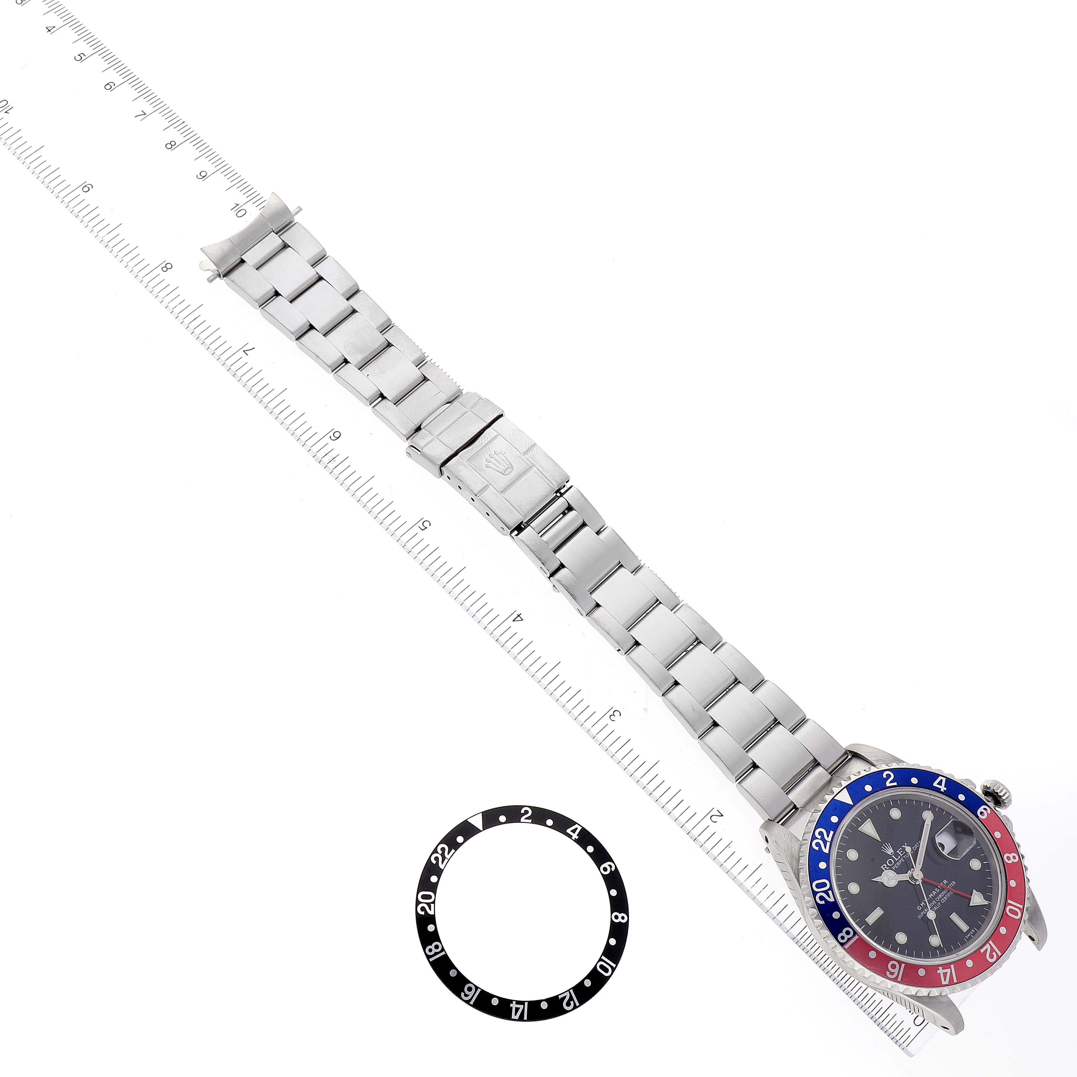 The image shows a top-down view of a Rolex GMT-Master 16700 Men
s Stainless Steel Black Dial 16700 Men
s Stainless Steel Black Dial watch with a metal bracelet and an extra bezel insert placed alongside a ruler.