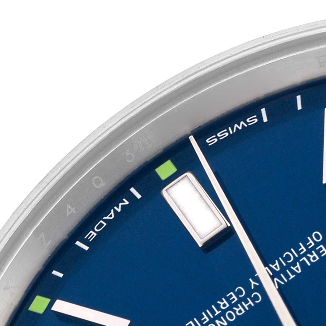 The image shows a close-up of the dial and bezel of a Rolex Oyster Perpetual 114300 Men's Stainless Steel Blue Dial 114300 Men's Stainless Steel Blue Dial watch, focusing on the indices and part of the branding.
