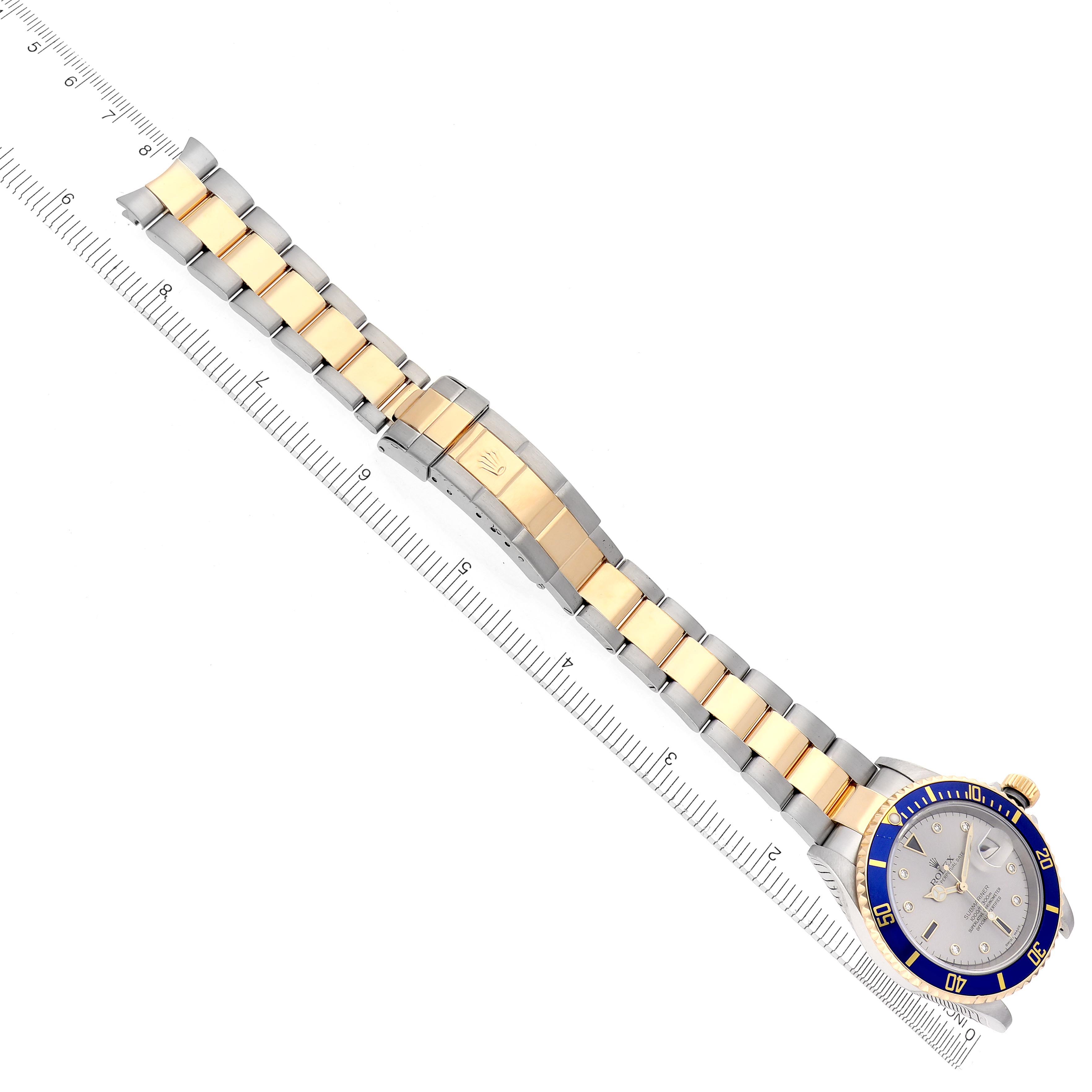 The Rolex Submariner 16613 Men's Steel and Gold (two tone) Silver Dial 16613 Men's Steel and Gold (two tone) Silver Dial is shown from an overhead angle, displaying its bezel, dial, and two-tone bracelet alongside a ruler.