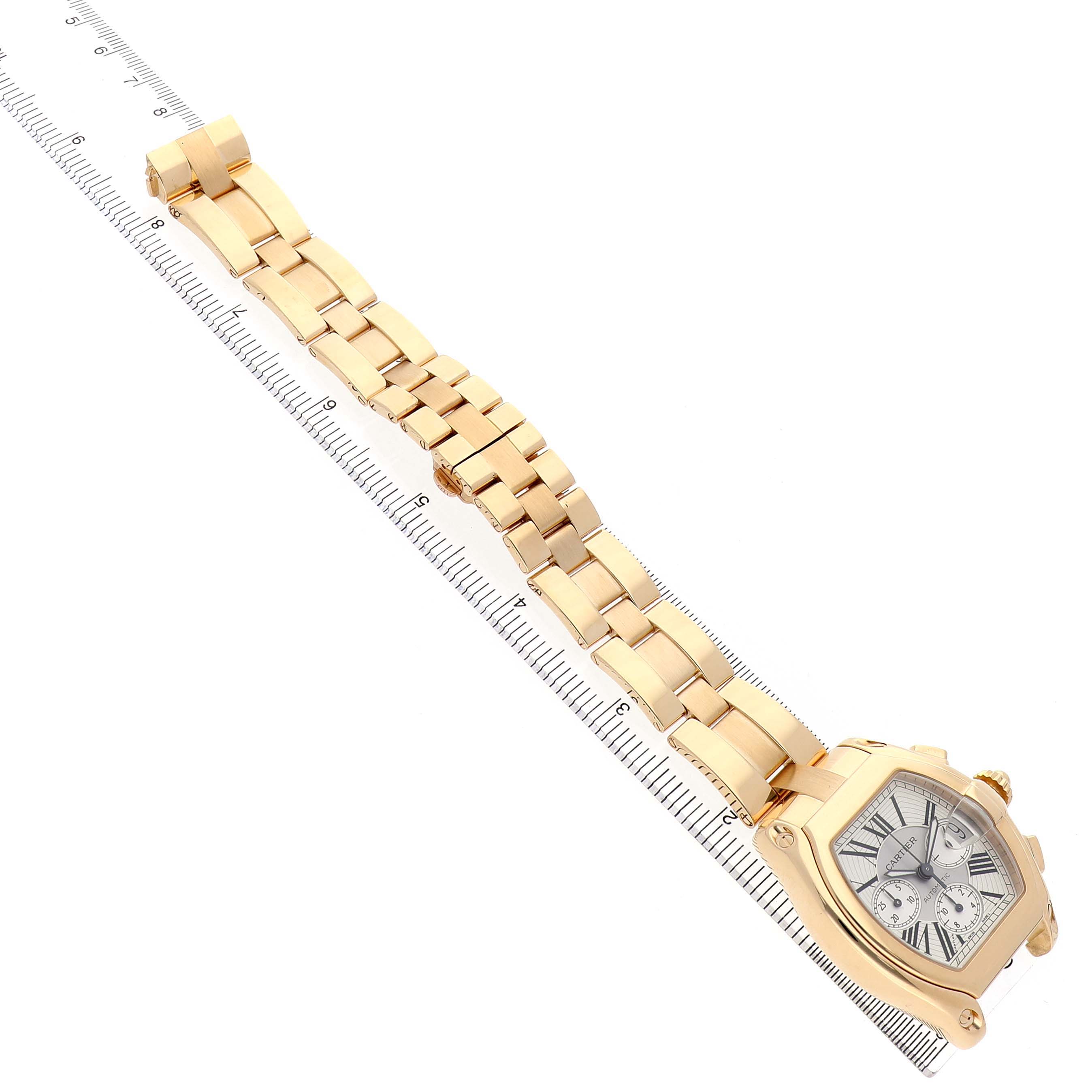 The Cartier Roadster W62021Y2 Men's Yellow Gold Silver Dial watch is shown at a slight side angle with focus on the dial and gold bracelet, placed diagonally against a ruler.