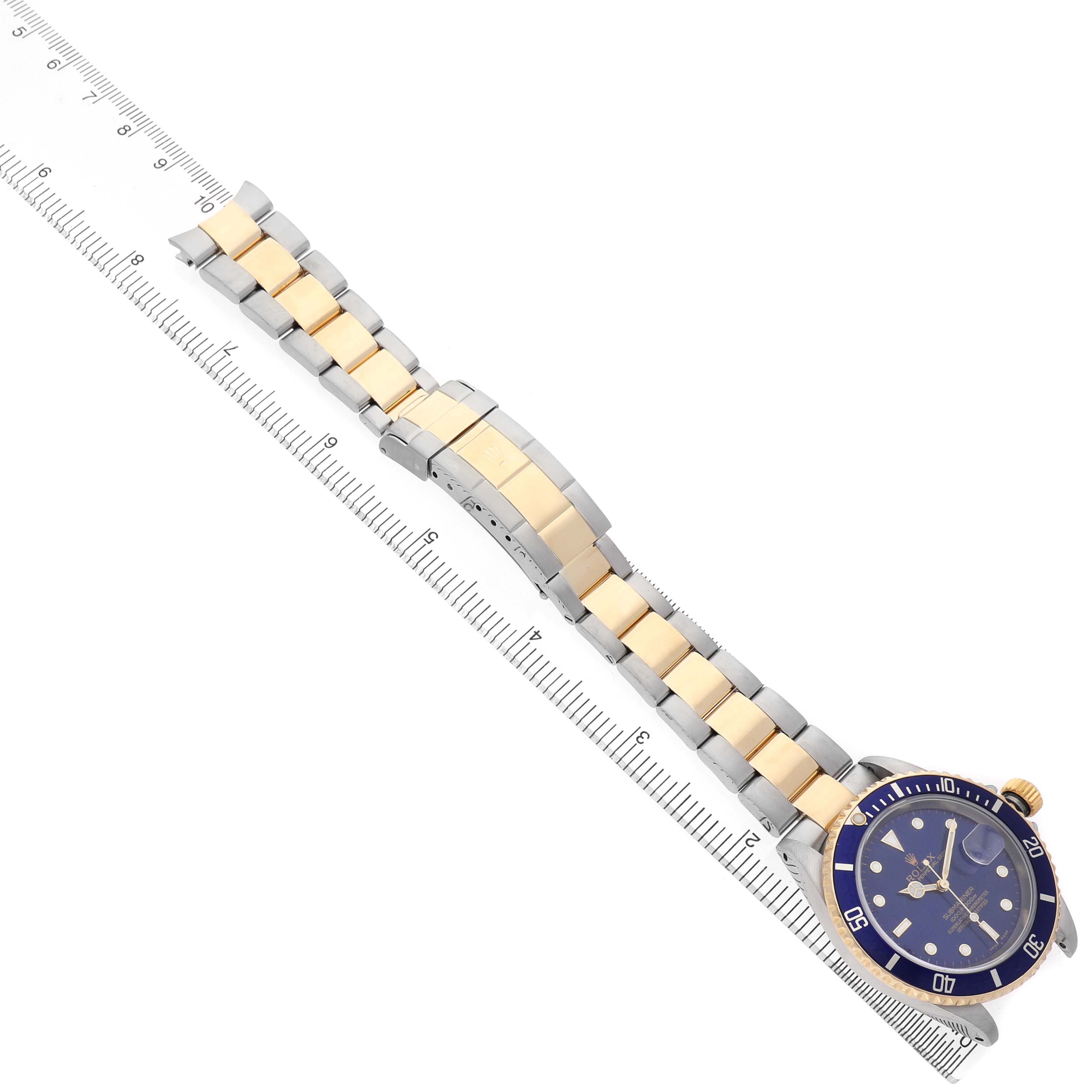 The Rolex Submariner 16613 Men's Steel and Gold (two tone) Blue Dial watch is shown from an overhead angle, featuring its blue dial, bezel, and two-tone bracelet alongside a ruler.