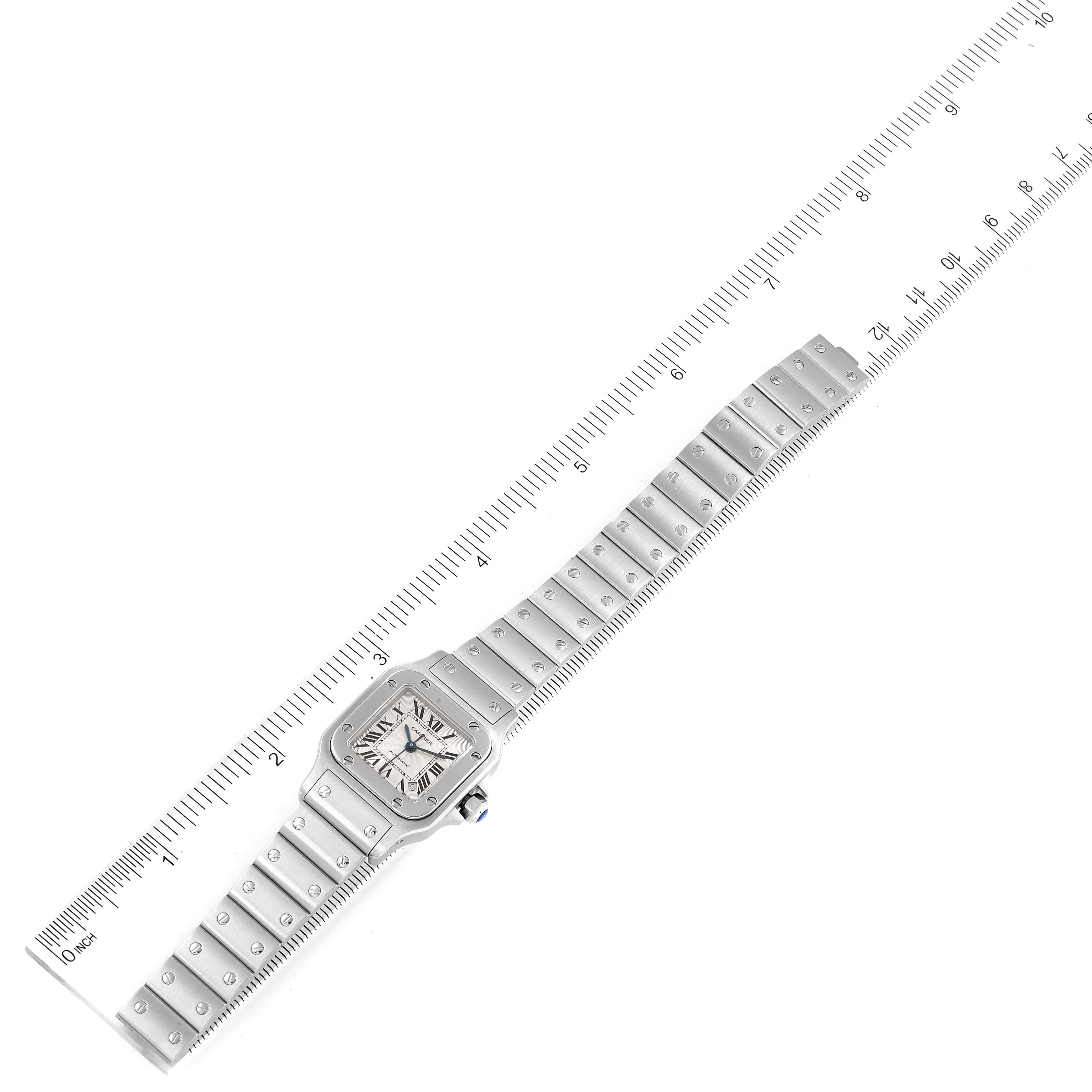 The image shows a Santos model watch by Cartier lying flat next to a ruler, displaying its face and stainless steel band.