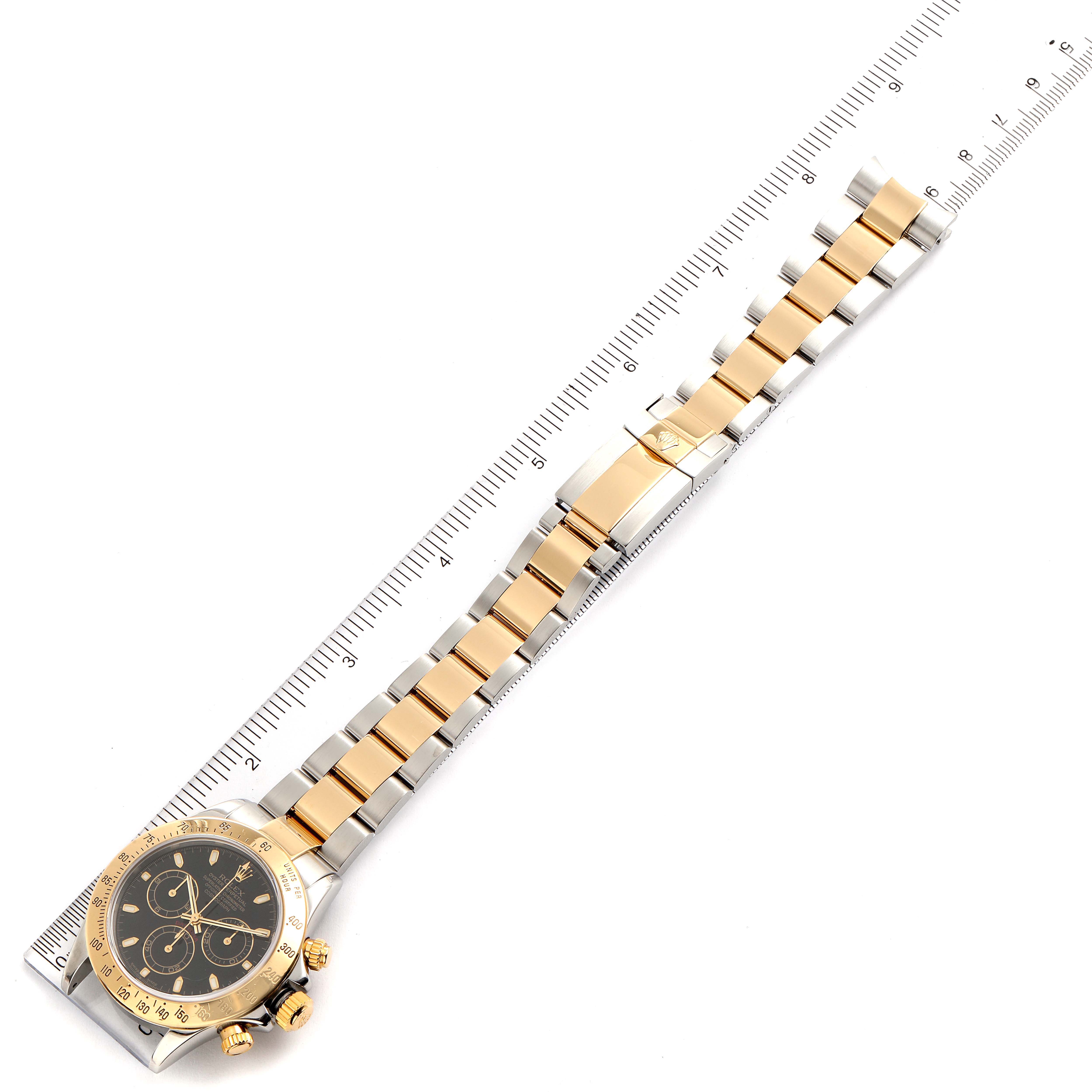 The image shows a Rolex Daytona 116523 Men's Steel and Gold (two tone) Black Dial 116523 Men's Steel and Gold (two tone) Black Dial watch laid flat, with a ruler underneath, displaying the face, bezel, bracelet, and clasp.