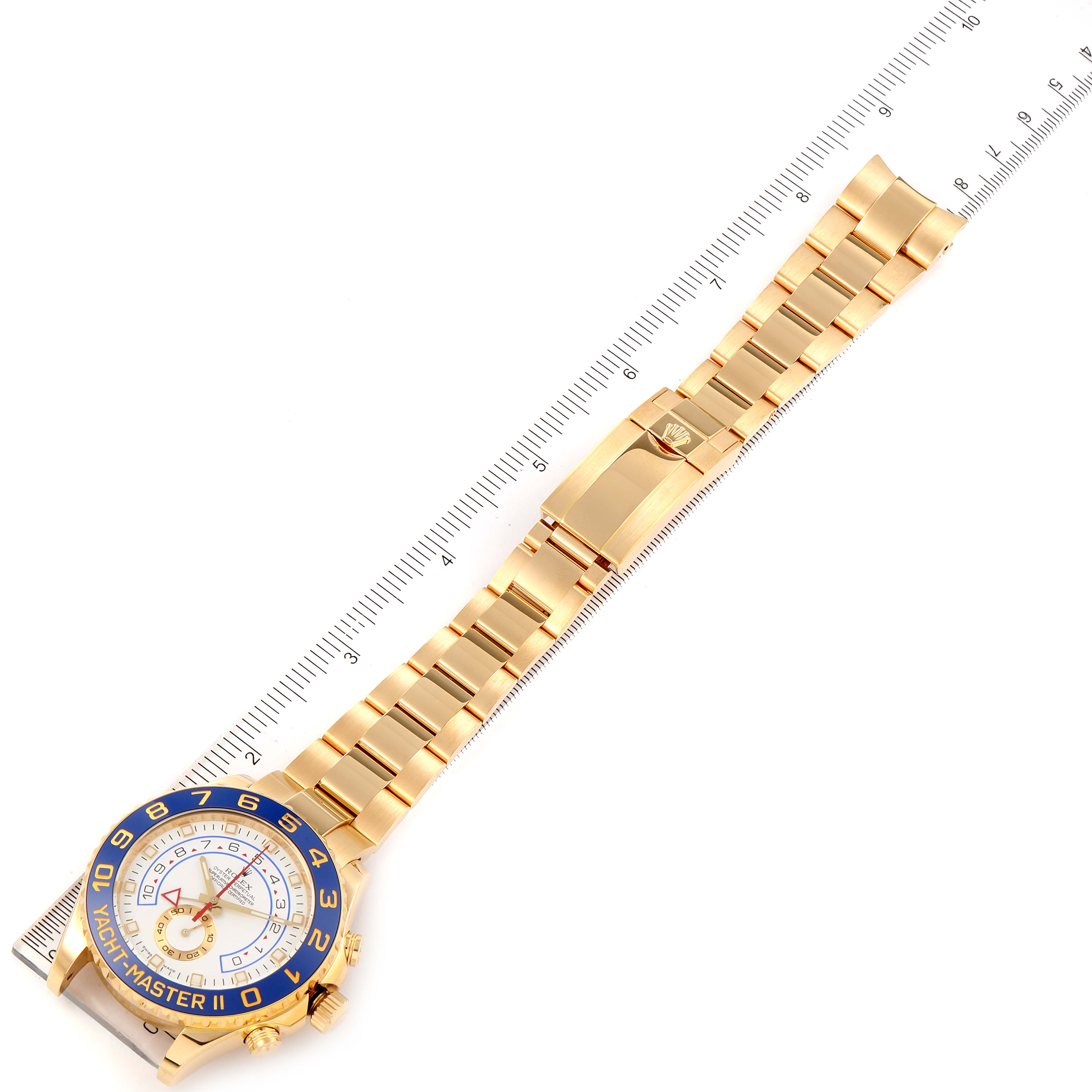 The Yacht-Master Rolex watch is shown from the top, displaying its gold bracelet, blue bezel, and clasp, measured against a ruler.