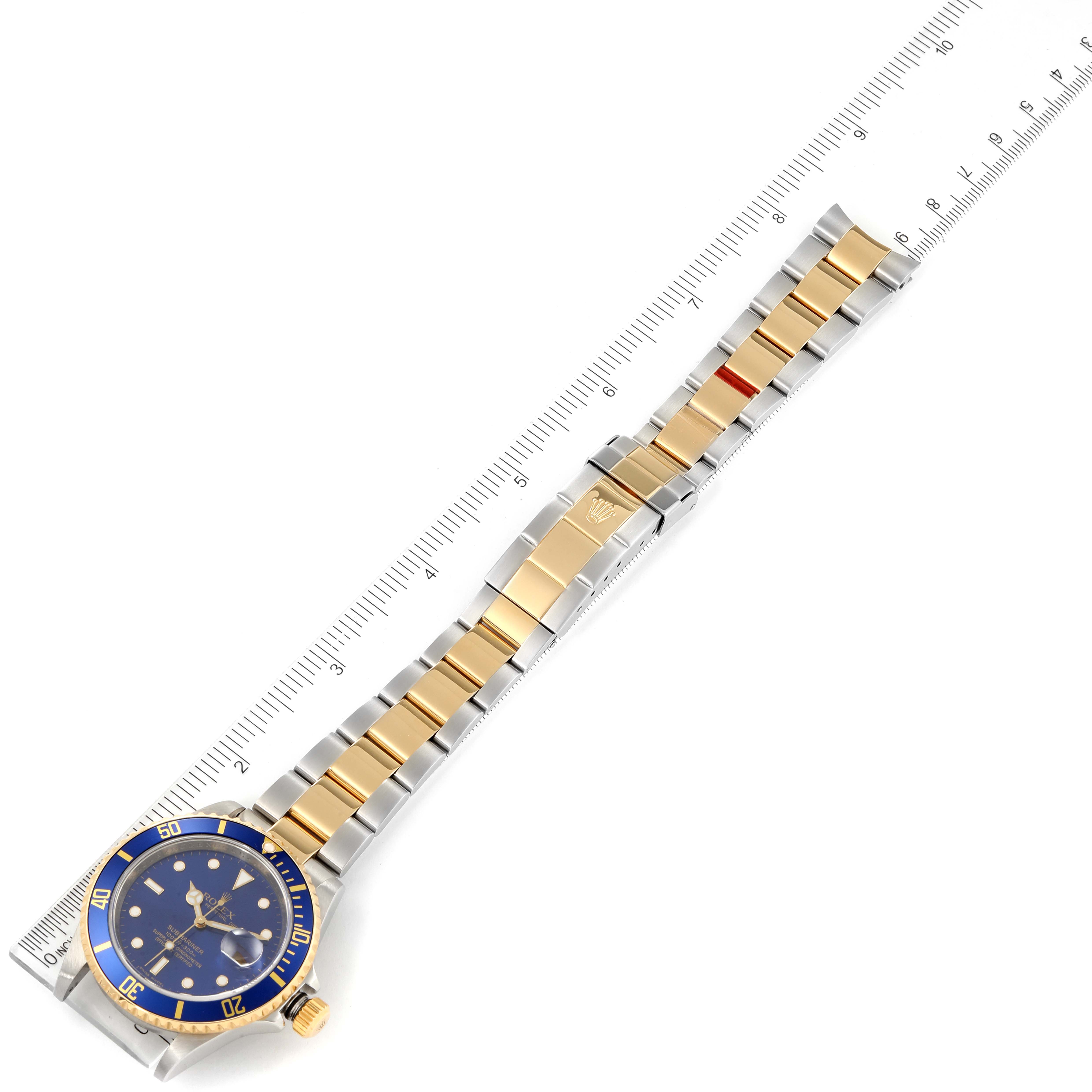 The image shows a Rolex Submariner 16613 Men
s Steel and Gold (two tone) Blue Dial 16613 Men
s Steel and Gold (two tone) Blue Dial watch with a blue dial and two-tone gold and silver bracelet, viewed from above with a ruler for scale.