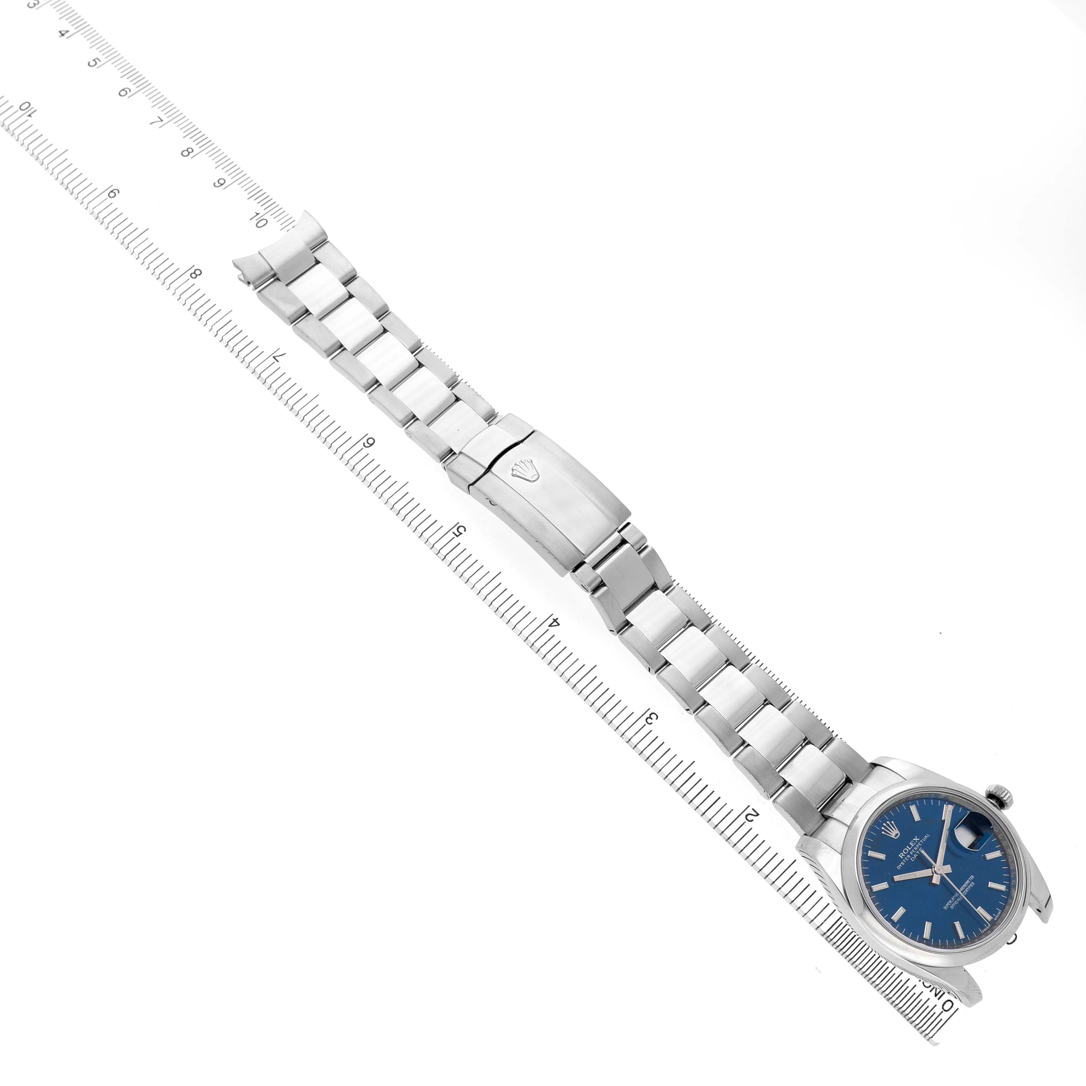 The Rolex Date 115200 Men's Stainless Steel Blue Dial model is shown at an angle displaying the band, clasp, and watch face, with a ruler underneath for scale.