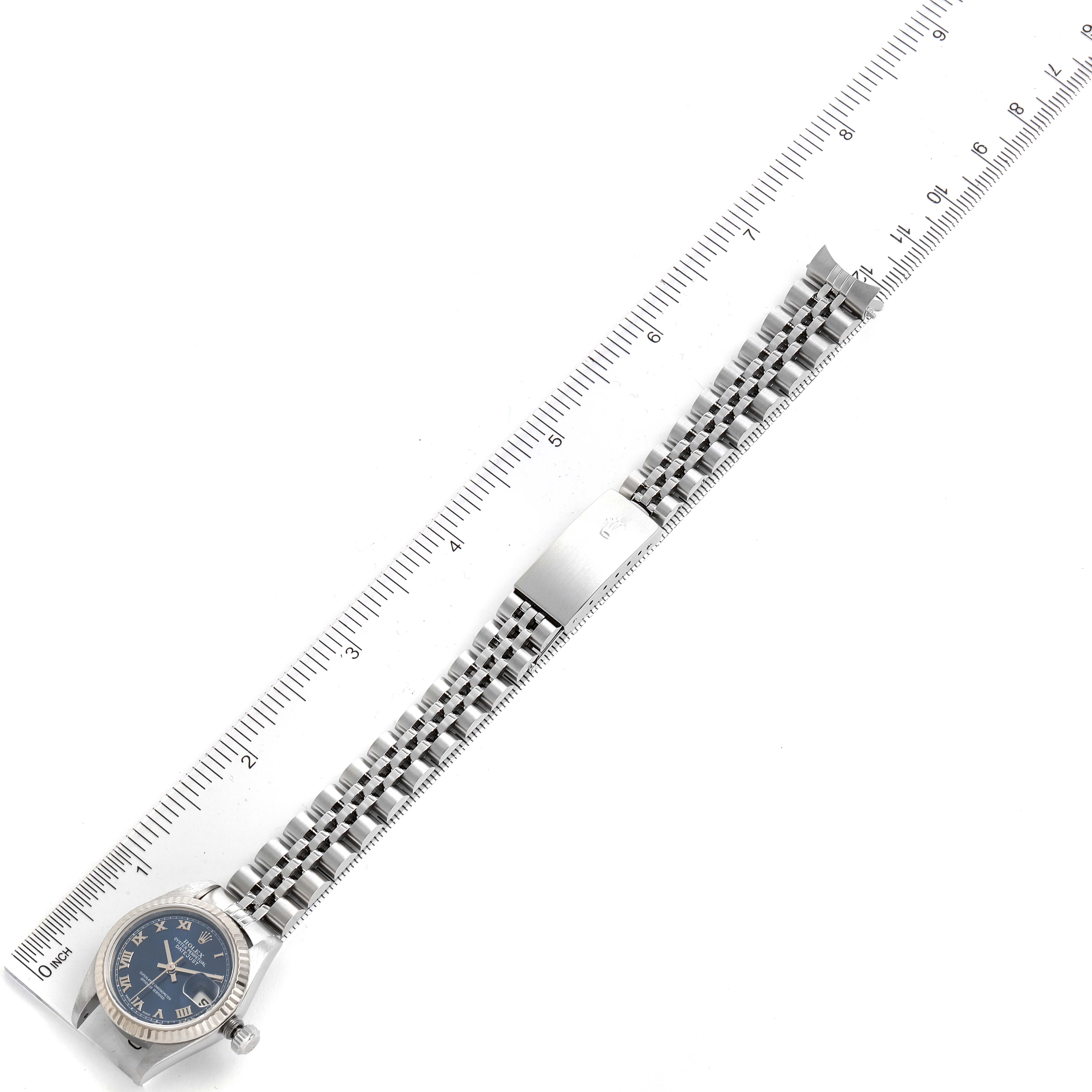 A Datejust model Rolex watch is shown from a top-down angle, highlighting the bracelet and clasp against a ruler background.
