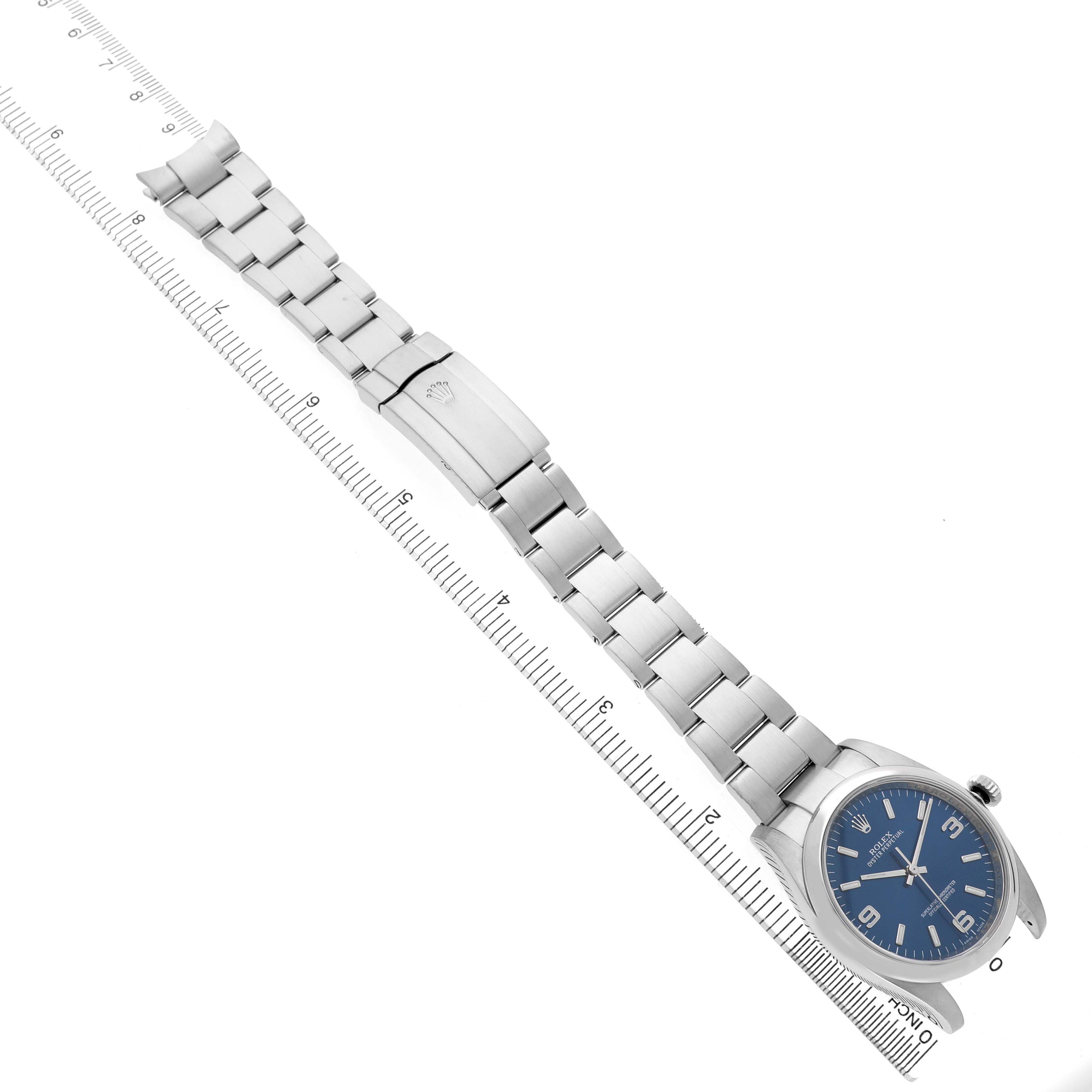 The Rolex Oyster Perpetual 116000 Men's Stainless Steel Blue Dial watch is displayed diagonally, showcasing the blue dial, bracelet, and clasp alongside a ruler.