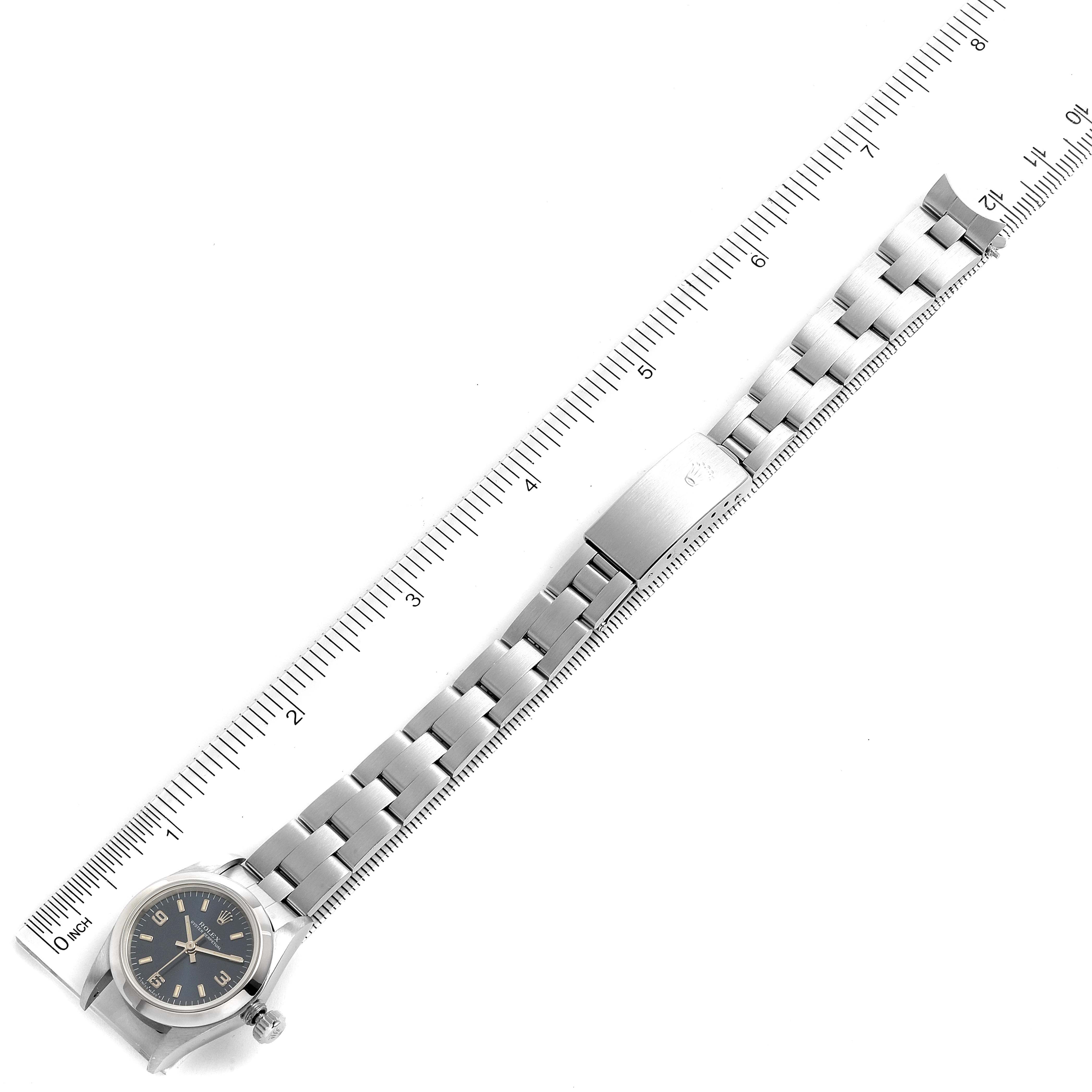 The image shows an overhead view of a Rolex Oyster Perpetual 67180 Women
s Stainless Steel Blue Dial 67180 Women
s Stainless Steel Blue Dial watch, including the face, bezel, crown, and entire metal bracelet alongside a ruler.