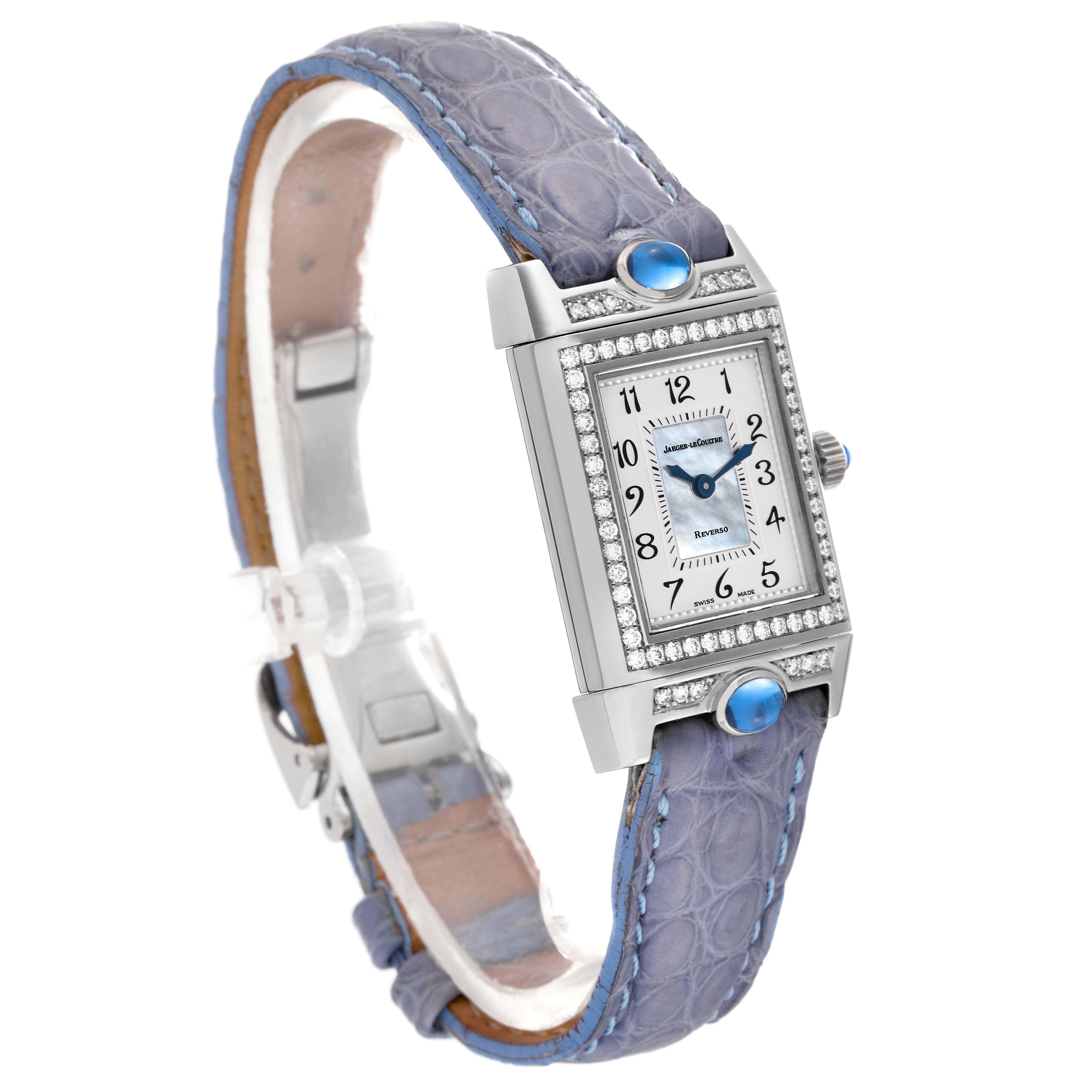The watch is a Jaeger LeCoultre Reverso Q2623403 Women's White Gold Mother of Pearl Dial Q2623403 Women's White Gold Mother of Pearl Dial model shown at an oblique angle, displaying its rectangular dial, blue leather strap, and bejeweled case.