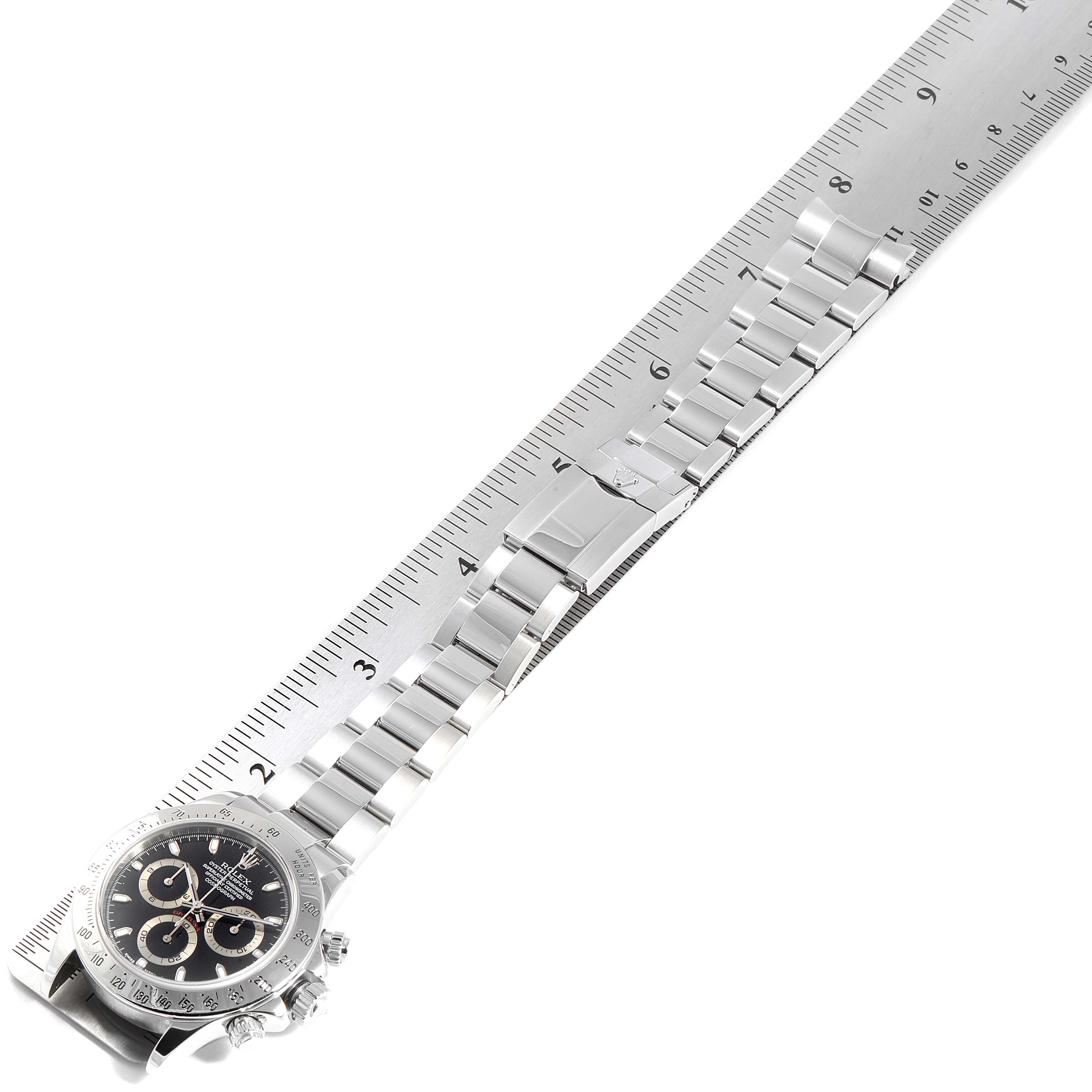 The image shows a Rolex Daytona 116520 Men's Stainless Steel Black Dial 116520 Men's Stainless Steel Black Dial watch placed flat along a ruler, displaying the dial, bezel, and full bracelet.