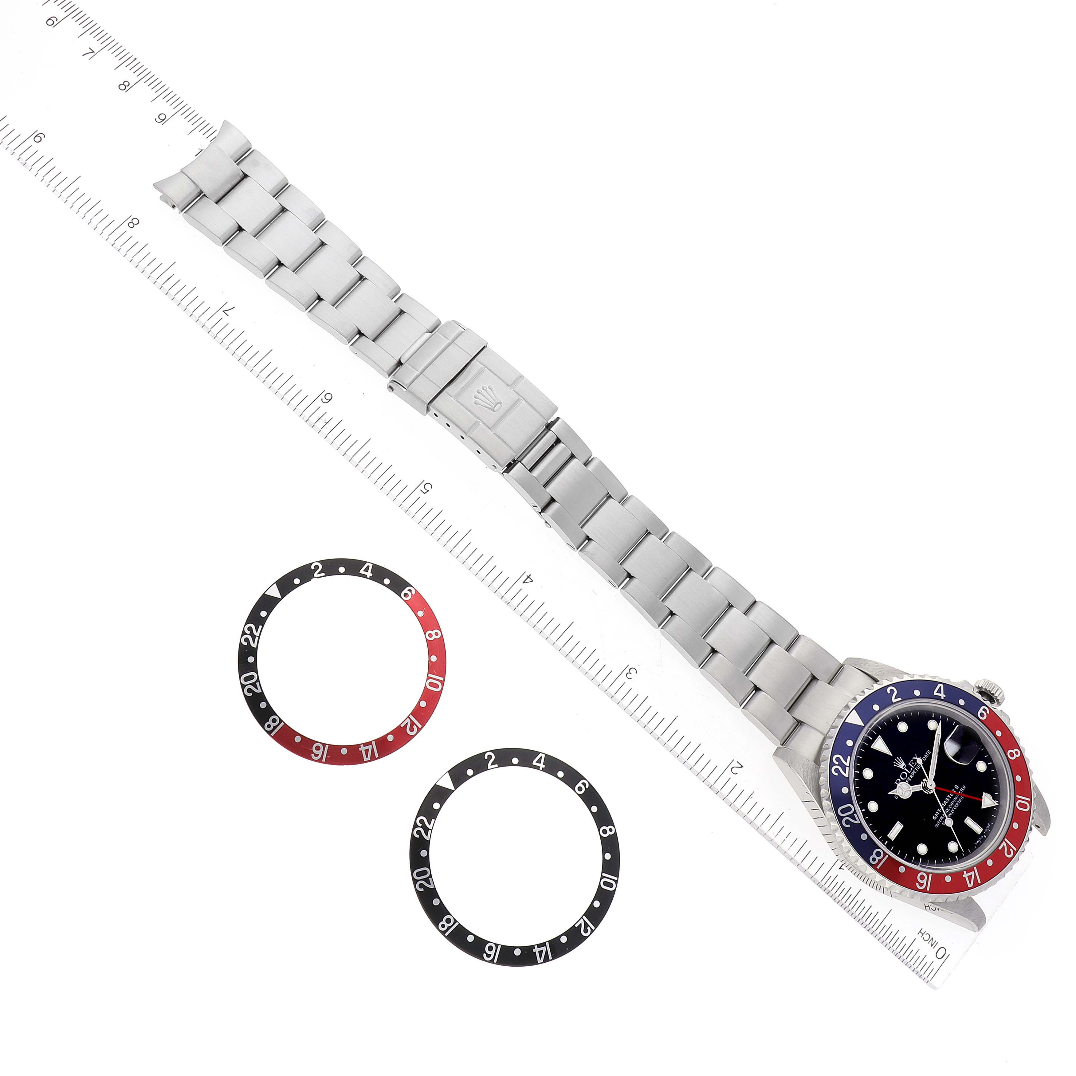 The image shows a top-down view of a Rolex GMT-Master 16710 Men
s Stainless Steel Black Dial 16710 Men
s Stainless Steel Black Dial watch and two separate bezel inserts along with a ruler.