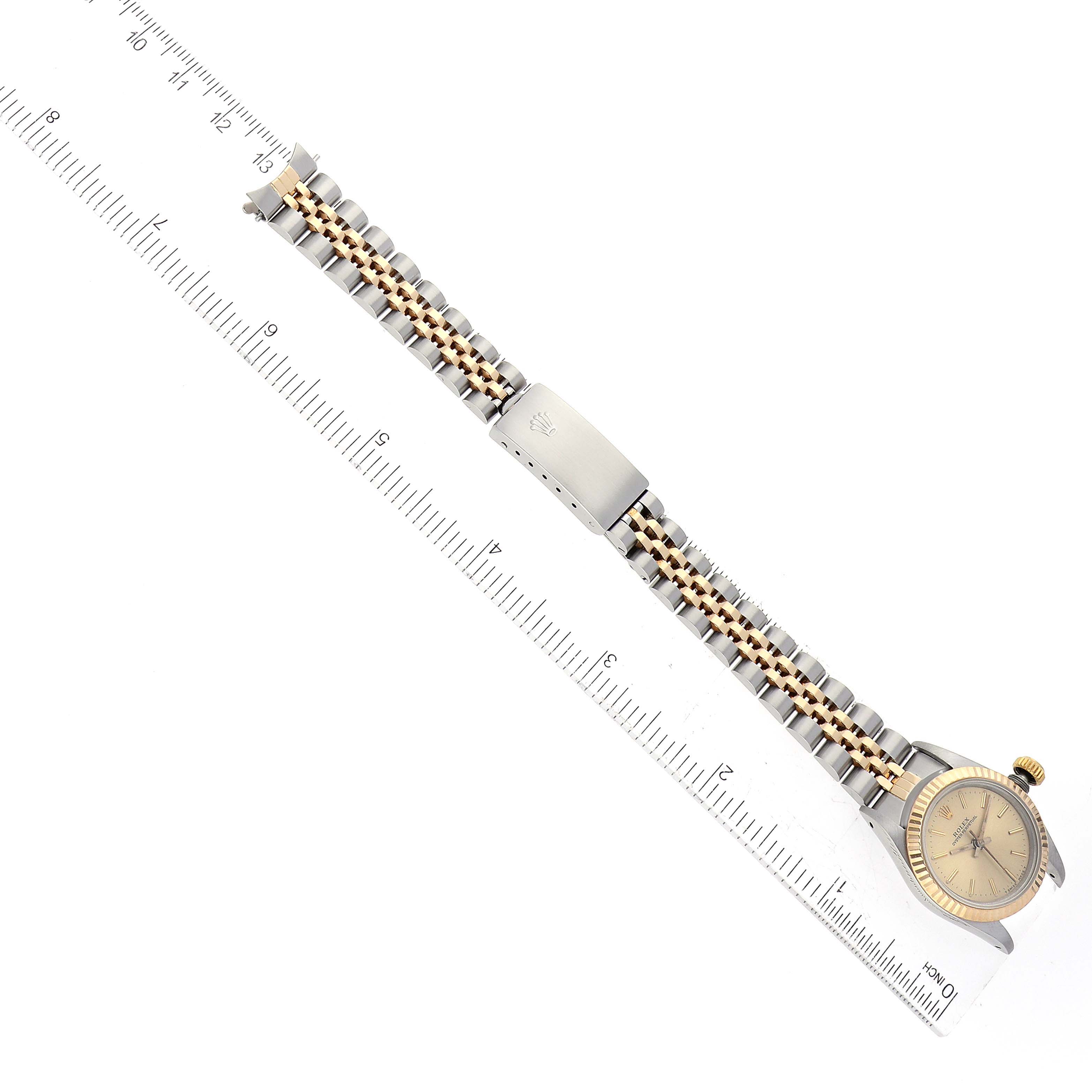 The image shows a top-down view of a Rolex Oyster Perpetual 67193 Women's Steel and Gold (two tone) Champagne Dial 67193 Women's Steel and Gold (two tone) Champagne Dial watch, including its face, bezel, and full bracelet alongside a ruler.
