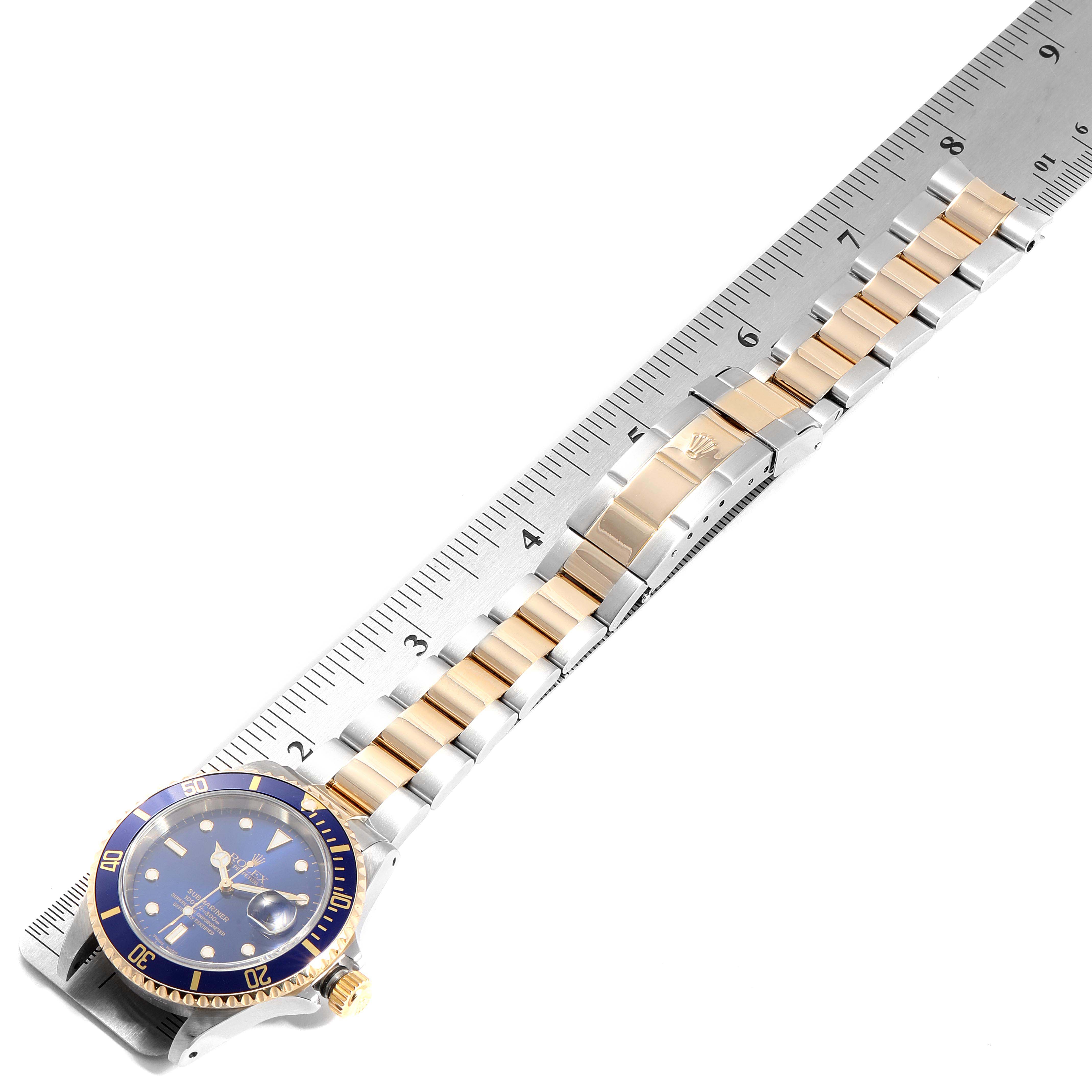 The image shows a Rolex Submariner 16613 Men
s Steel and Gold (two tone) Blue Dial 16613 Men
s Steel and Gold (two tone) Blue Dial watch laid flat on a ruler, displaying its full band and face.