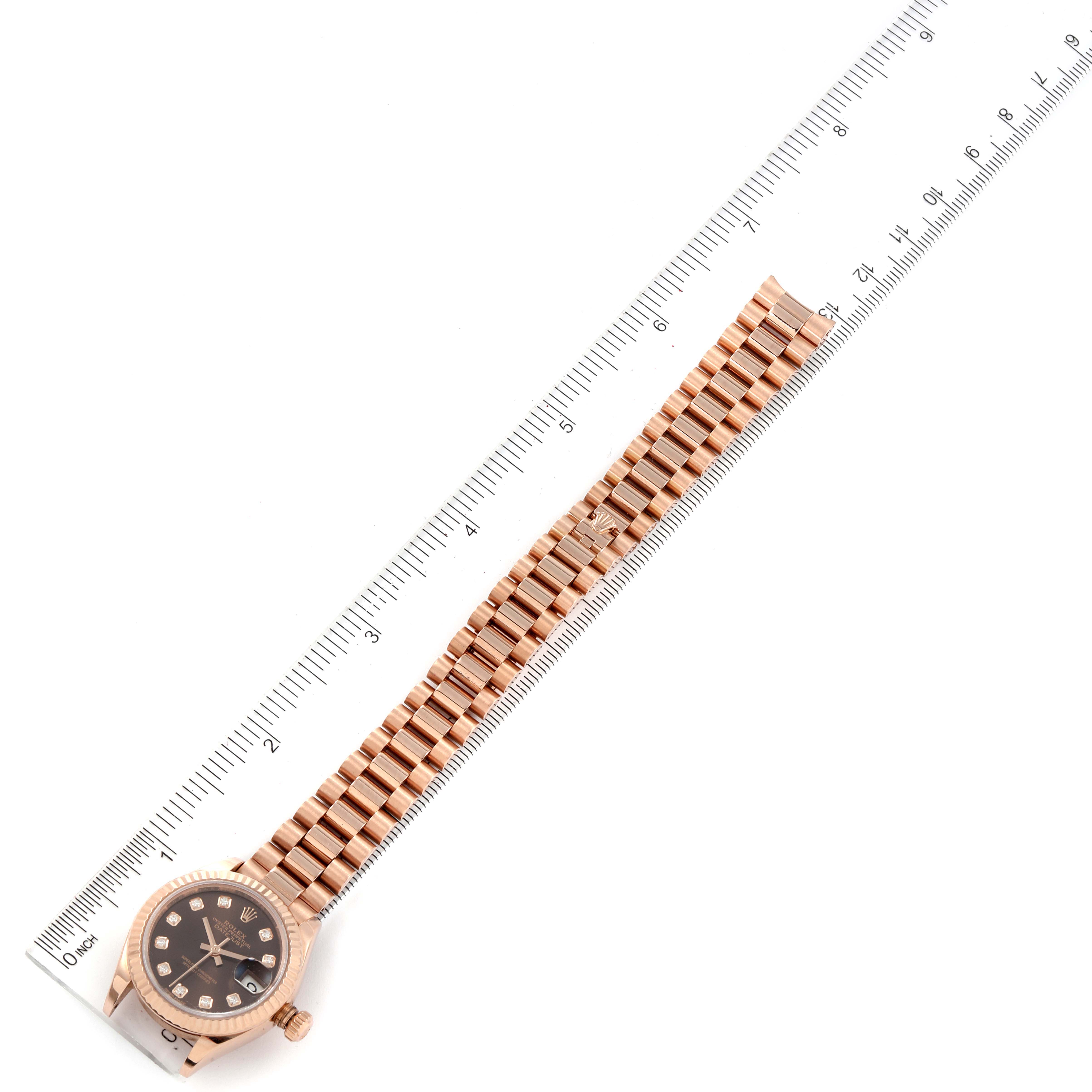 The Rolex President 279175 Women
s Rose Gold Brown Dial 279175 Women
s Rose Gold Brown Dial model watch is shown from a top view, displaying its face, crown, and full metal bracelet alongside a ruler.