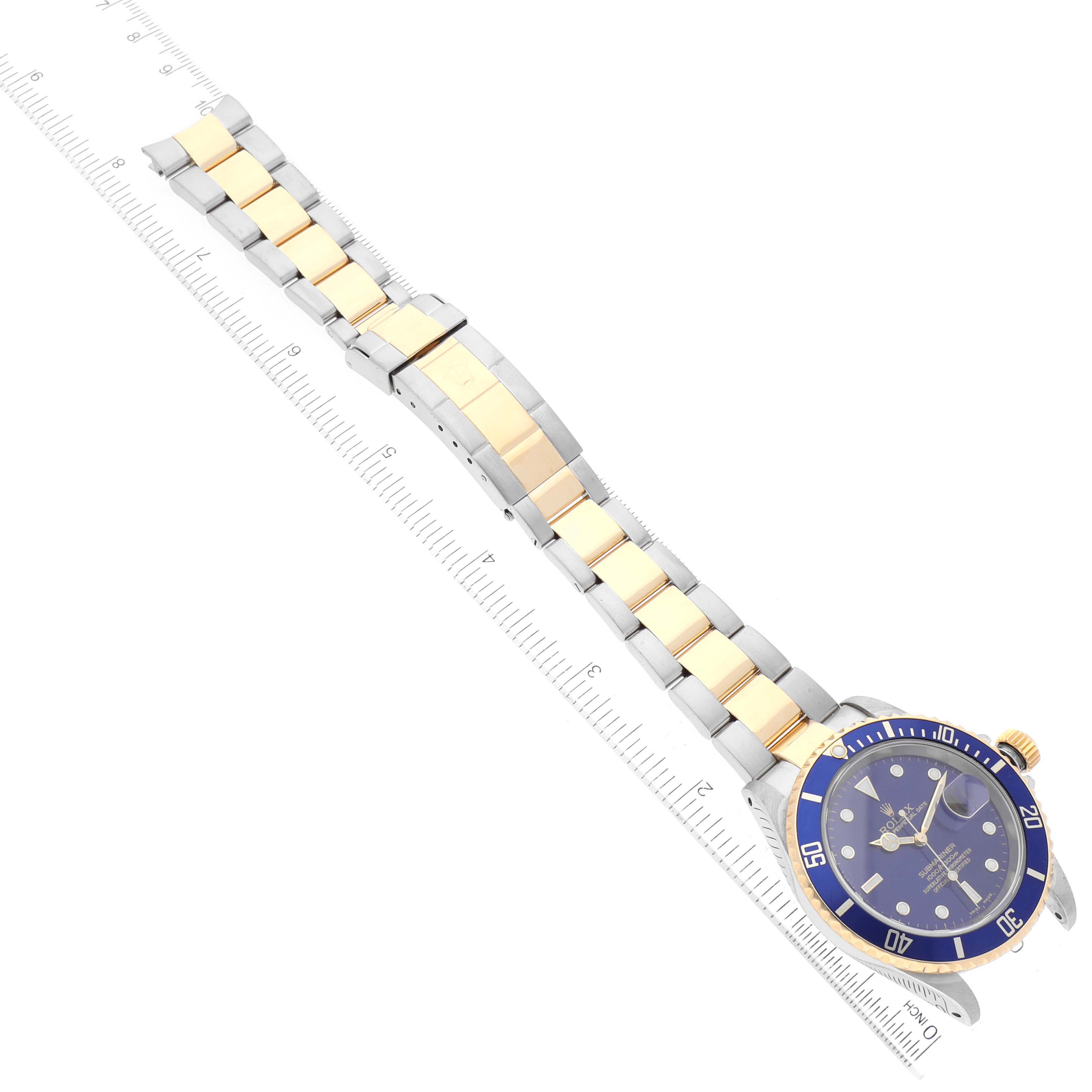 The image shows an overhead view of a Rolex Submariner 16613 Men's Steel and Gold (two tone) Blue Dial 16613 Men's Steel and Gold (two tone) Blue Dial watch, featuring a blue dial, bezel, and a two-tone gold and silver bracelet, with a ruler for scale.