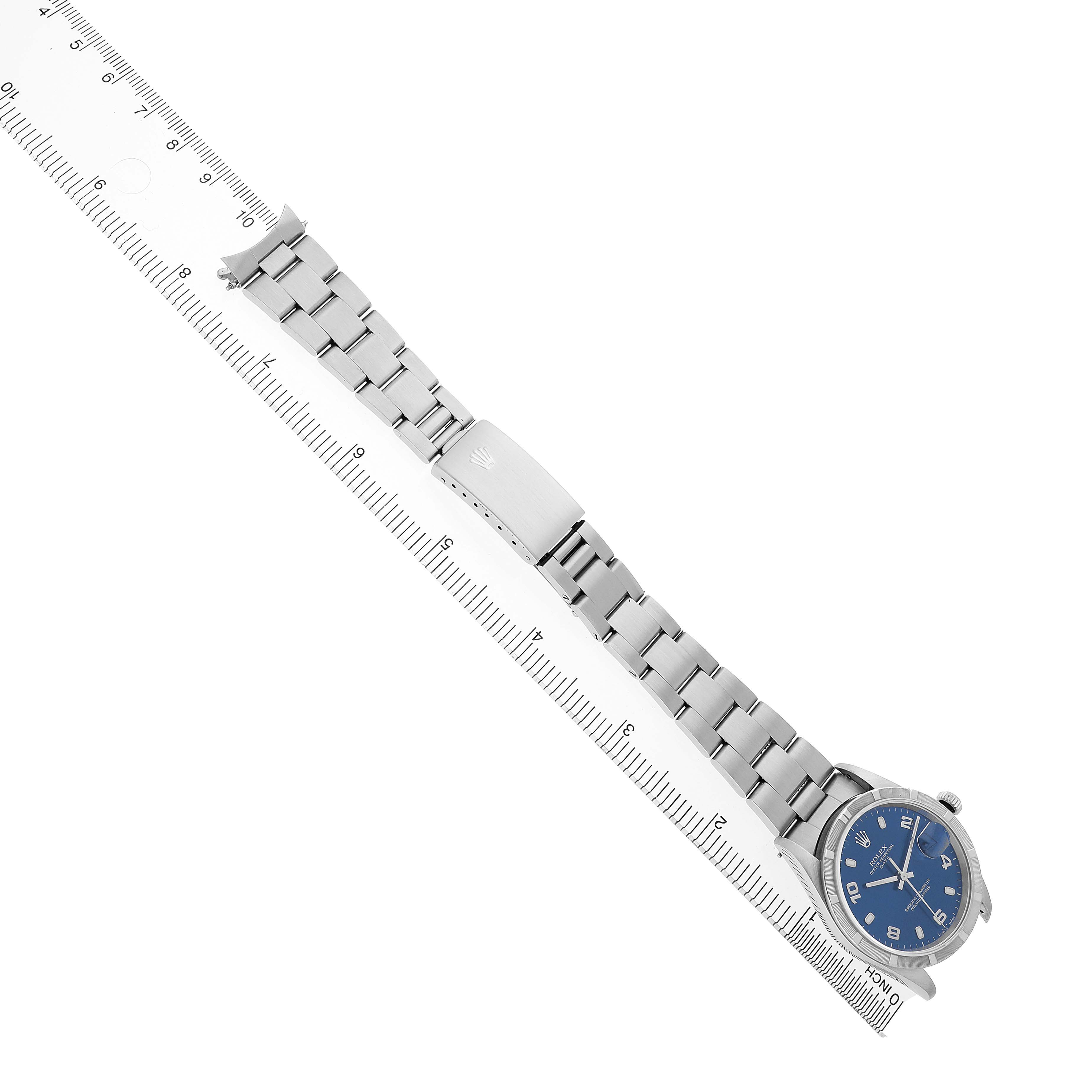 The Rolex Date 15210 Men's Stainless Steel Blue Dial model is shown at a diagonal angle, featuring the watch face, clasp, and bracelet aligned with a ruler.