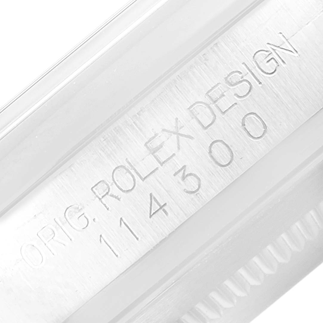 The image shows a close-up of the side of a Rolex Oyster Perpetual 114300 Men's Stainless Steel Silver Dial 114300 Men's Stainless Steel Silver Dial watch, highlighting the engraved "ORIG. ROLEX DESIGN 114300."