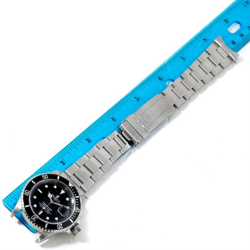 The image shows a Rolex Submariner 16610 Men
s Stainless Steel Black Dial 16610 Men
s Stainless Steel Black Dial watch positioned next to a ruler, highlighting its face, bezel, bracelet, and clasp.