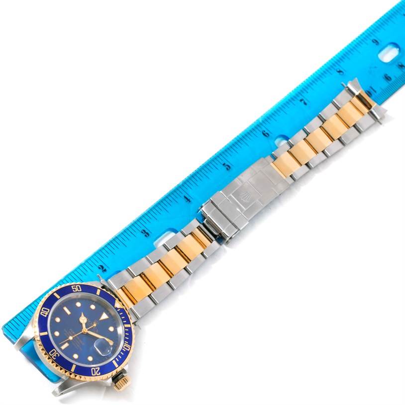 The image shows a Rolex Submariner 16613 Men's Steel and Gold (two tone) Blue Dial 16613 Men's Steel and Gold (two tone) Blue Dial watch placed on a ruler, displaying its blue dial, bezel, and two-tone bracelet.