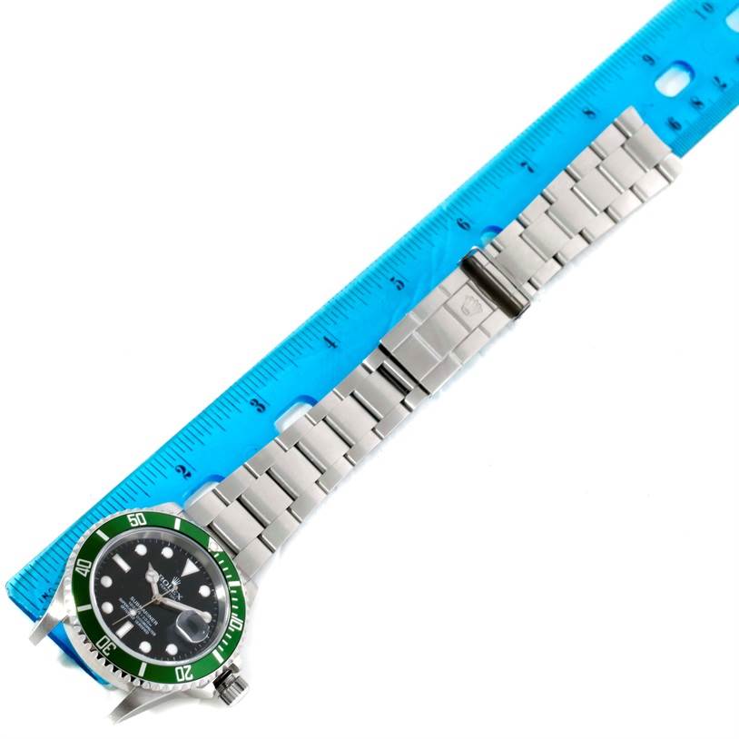 The image shows a Rolex Submariner 16610LV Men
s Stainless Steel Black Dial 16610LV Men
s Stainless Steel Black Dial watch lying flat with a green bezel and stainless steel bracelet next to a blue ruler.
