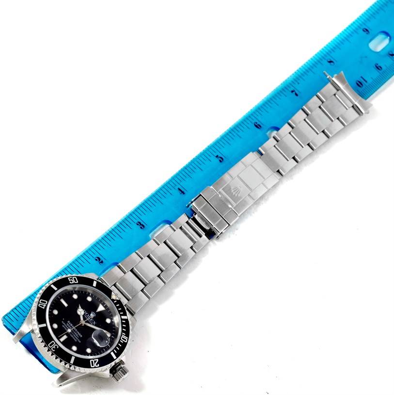 This image shows a Rolex Submariner 16610 Men
s Stainless Steel Black Dial 16610 Men
s Stainless Steel Black Dial watch laid flat against a blue ruler, displaying the watch face, bracelet, and clasp.
