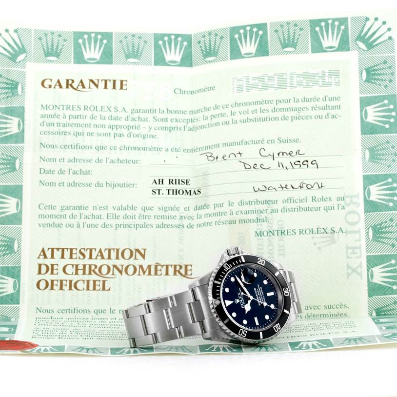 The image shows a Rolex Submariner 16610 Men
s Stainless Steel Black Dial 16610 Men
s Stainless Steel Black Dial watch positioned on top of its warranty certificate and chronometer attestation.