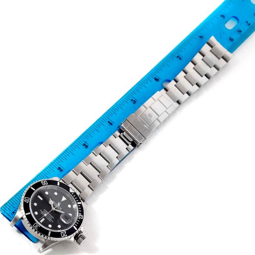 The Rolex Submariner 16610 Men
s Stainless Steel Black Dial 16610 Men
s Stainless Steel Black Dial watch is displayed flat with its stainless steel bracelet extended and measured against a blue ruler.