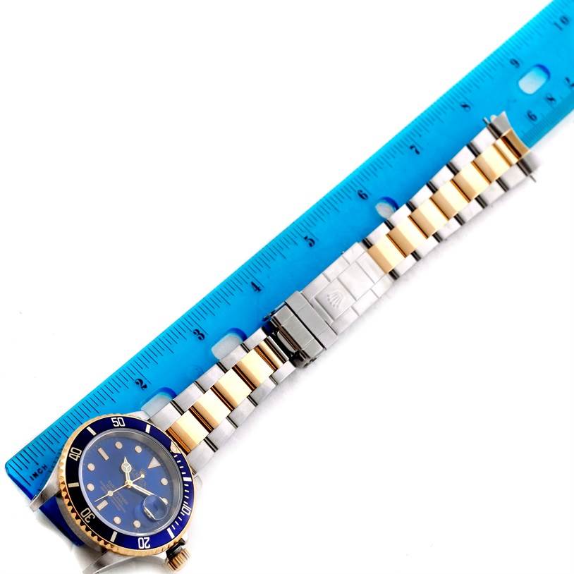 The image shows the Rolex Submariner 16613 Men
s Steel and Gold (two tone) Blue Dial 16613 Men
s Steel and Gold (two tone) Blue Dial watch face, crown, and bracelet, measured against a blue ruler from an overhead angle.
