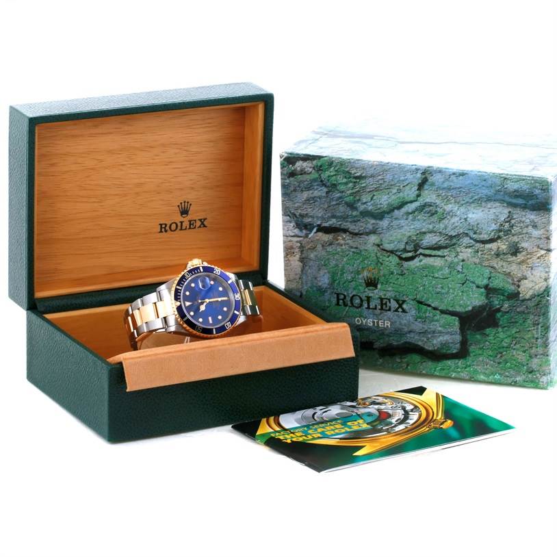 The Rolex Submariner 16613 Men
s Steel and Gold (two tone) Blue Dial 16613 Men
s Steel and Gold (two tone) Blue Dial watch is shown in its open box at an angled view with its packaging and booklet nearby.