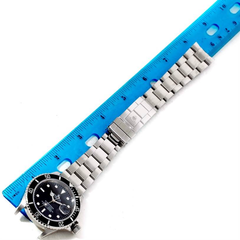 This image shows a Rolex Submariner 16610 Men
s Stainless Steel Black Dial 16610 Men
s Stainless Steel Black Dial watch laid flat with a blue ruler placed alongside the metal bracelet.