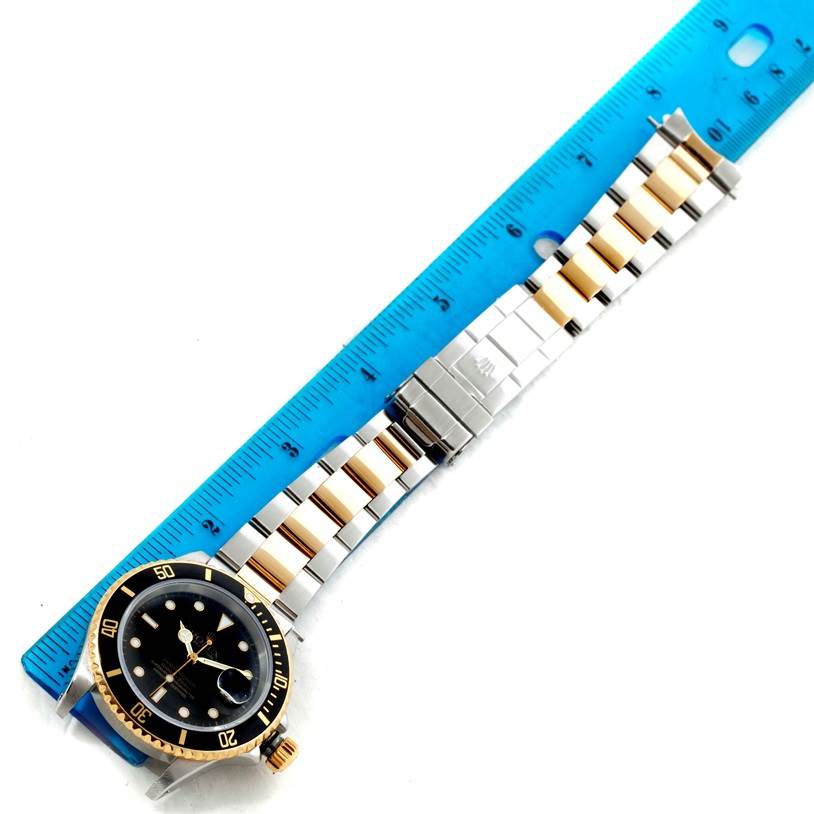 The image shows a Rolex Submariner 16613 Men's Steel and Gold (two tone) Black Dial 16613 Men's Steel and Gold (two tone) Black Dial laid flat next to a blue ruler for scale.