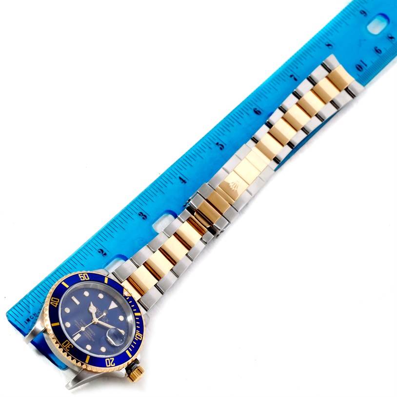 The Rolex Submariner 16613 Men
s Steel and Gold (two tone) Blue Dial 16613 Men
s Steel and Gold (two tone) Blue Dial watch is shown from a top-down angle, displaying the face, bezel, and two-tone metal band alongside a blue ruler.