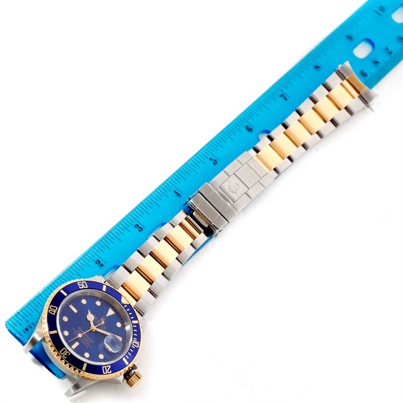 The image shows a Rolex Submariner 16613 Men
s Steel and Gold (two tone) Blue Dial 16613 Men
s Steel and Gold (two tone) Blue Dial watch lying flat with its blue dial, bezel, and two-tone metal bracelet fully visible alongside a blue ruler.