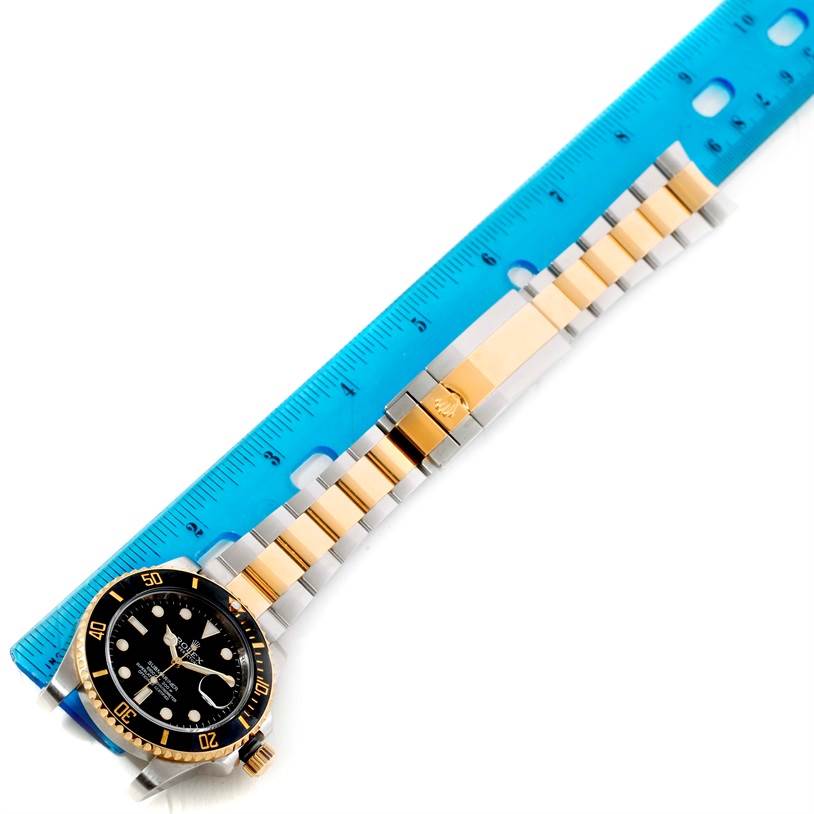 The image shows a Rolex Submariner 116613 Men
s Steel and Gold (two tone) Black Dial 116613 Men
s Steel and Gold (two tone) Black Dial watch, displaying the face and linked metal strap, next to a blue ruler.