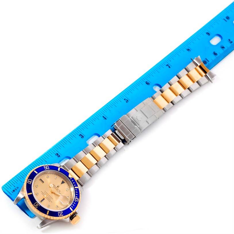 The image shows a Rolex Submariner 16613 Men's Steel and Gold (two tone) Champagne Dial 16613 Men's Steel and Gold (two tone) Champagne Dial watch laid flat next to a blue ruler, focusing on the watch face and metal band.