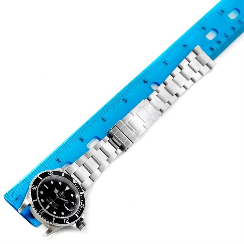 The Rolex Submariner 16610 Men
s Stainless Steel Black Dial 16610 Men
s Stainless Steel Black Dial watch is shown from a top-down angle with its full bracelet extended alongside a blue ruler.