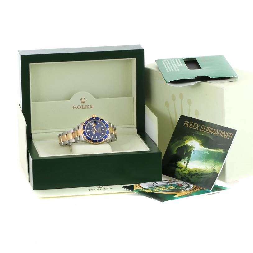 The Rolex Submariner 16613 Men's Steel and Gold (two tone) Blue Dial 16613 Men's Steel and Gold (two tone) Blue Dial watch is displayed front-facing inside its open box, with accompanying booklets and accessories nearby.