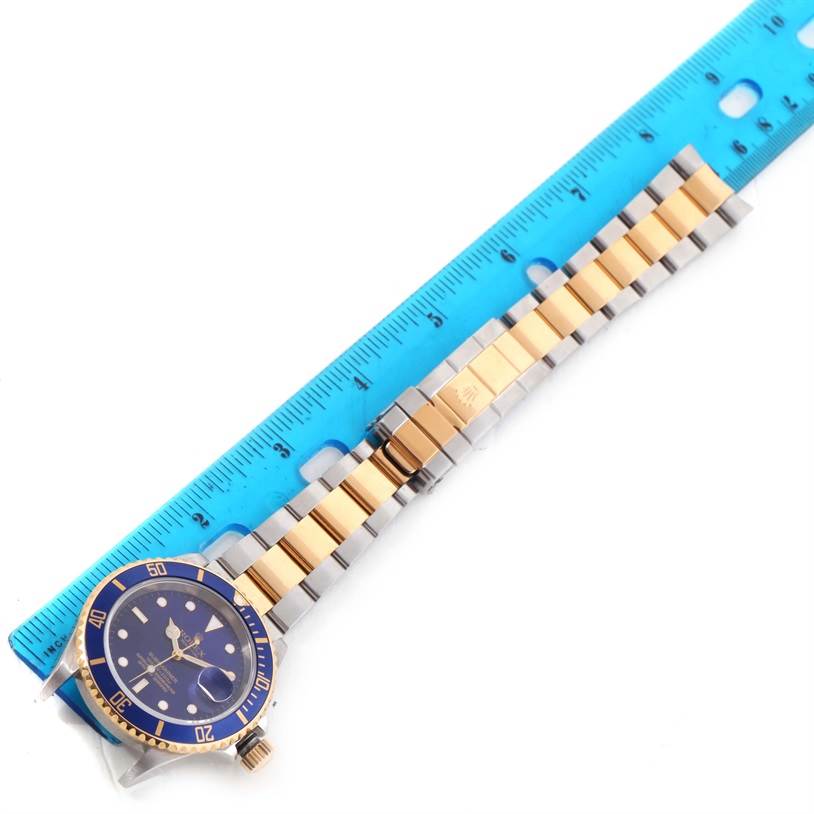 This image shows a Rolex Submariner 16613 Men's Steel and Gold (two tone) Blue Dial 16613 Men's Steel and Gold (two tone) Blue Dial watch flat with its blue bezel, dial, and two-tone bracelet next to a ruler.