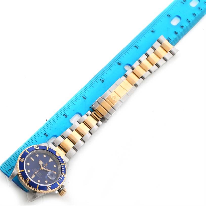 The Rolex Submariner 16613 Men's Steel and Gold (two tone) Blue Dial 16613 Men's Steel and Gold (two tone) Blue Dial watch is shown from above, including the face, bezel, and the two-tone bracelet with a blue ruler beside it.