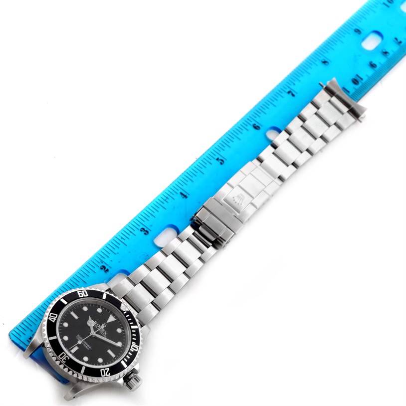 The image shows a Rolex Submariner 14060 Men
s Stainless Steel Black Dial 14060 Men
s Stainless Steel Black Dial watch laid flat next to a blue ruler, highlighting the watch body, bracelet, and clasp.