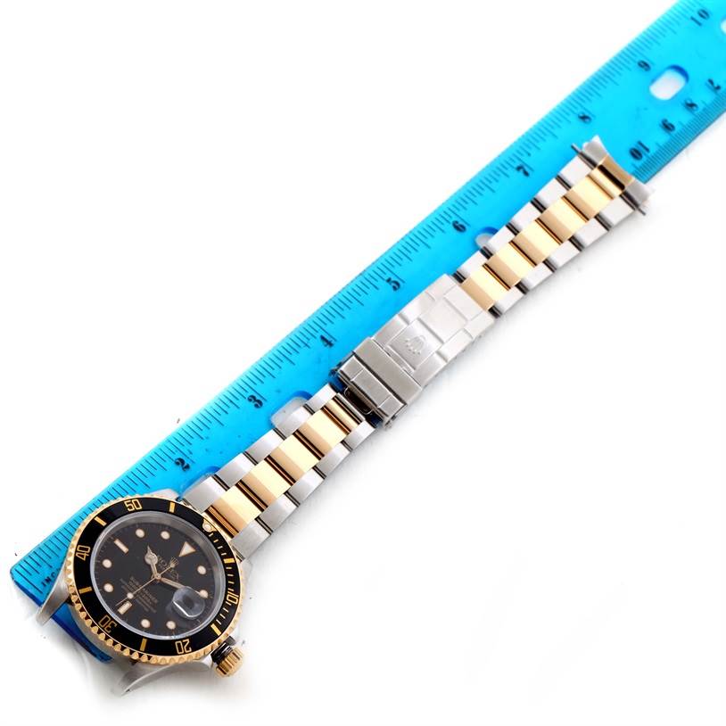 The image shows a top-down view of a Rolex Submariner 16613 Men's Steel and Gold (two tone) Black Dial 16613 Men's Steel and Gold (two tone) Black Dial watch alongside a blue measuring ruler.