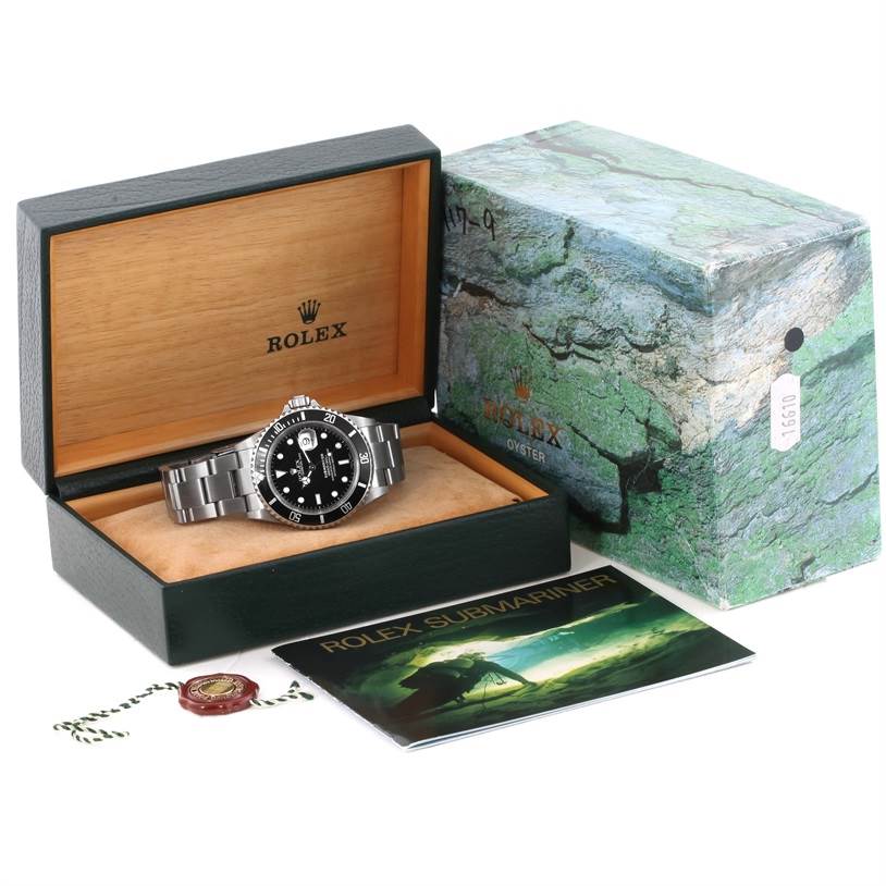 The image shows a Rolex Submariner 16610 Men
s Stainless Steel Black Dial 16610 Men
s Stainless Steel Black Dial watch from an angled view inside an open box, alongside its green outer box, manual, and tags.